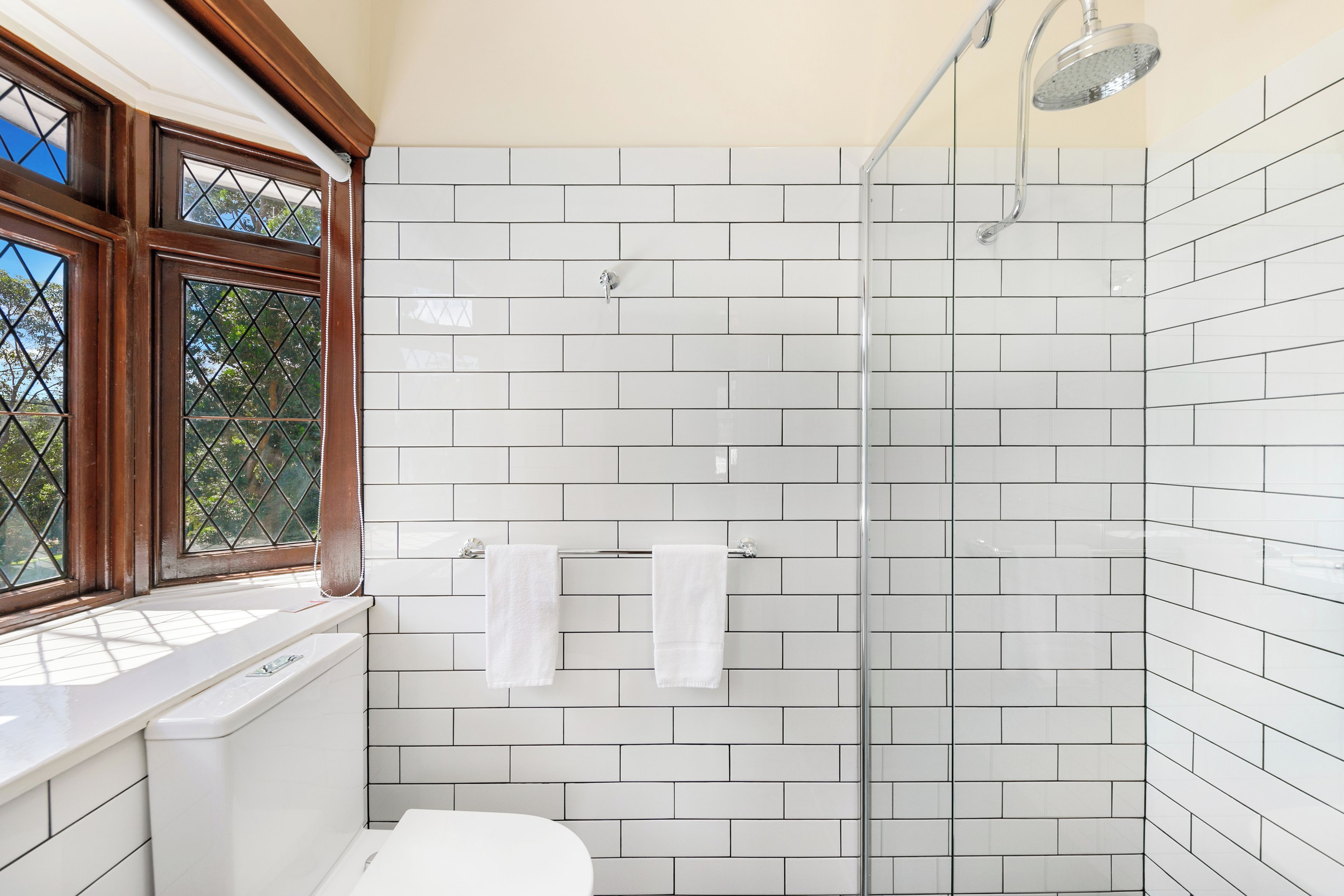 A bathroom with white tiled walls, a shower, and a window with a view of trees.