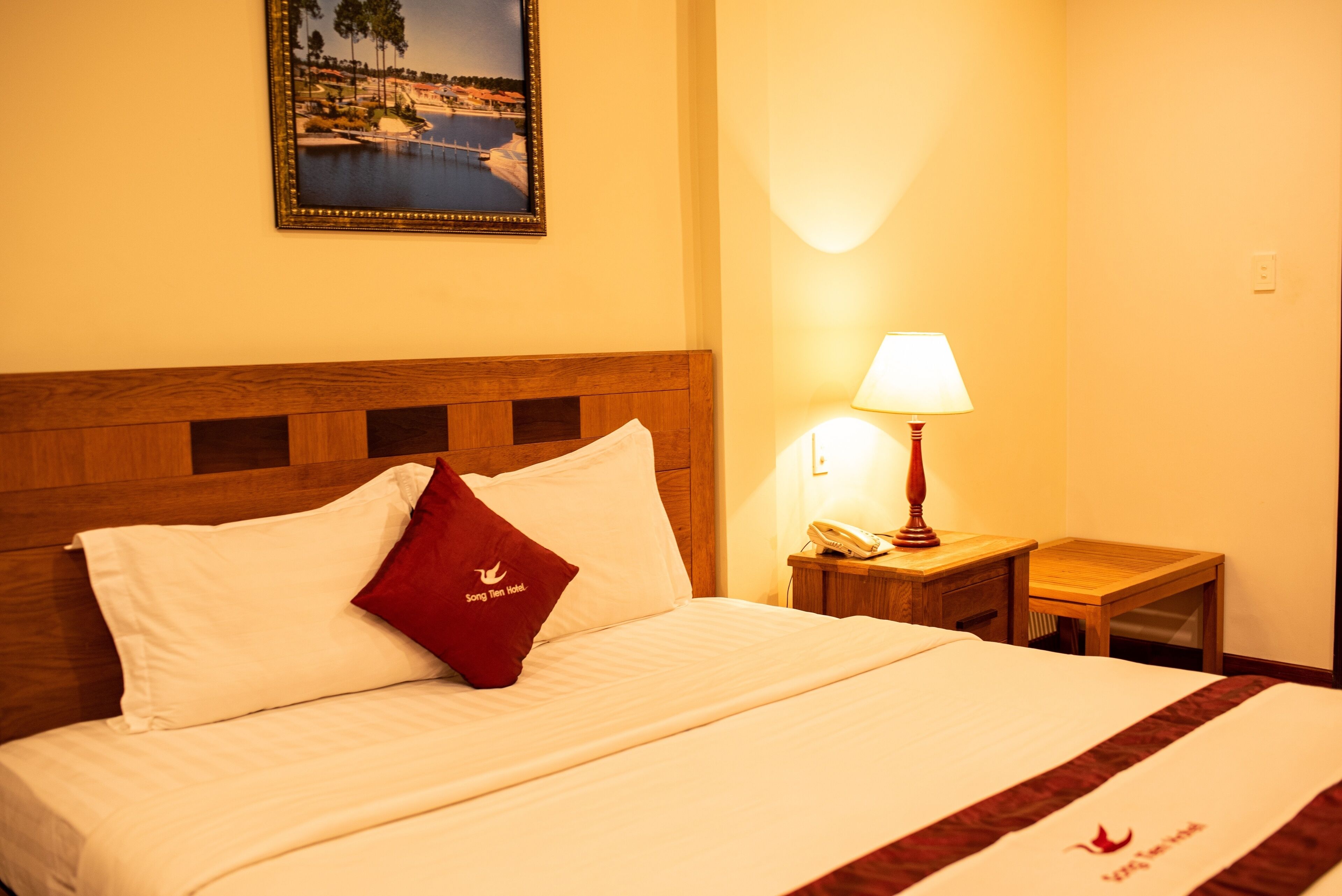 Quarto de hotel com uma cama bem arrumada, cabeceira de madeira, mesa de cabeceira com abajur e um quadro na parede.