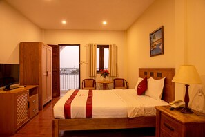 A hotel room with a bed, a desk with a lamp, a TV, a wardrobe, a window with curtains, a small table with flowers, and a painting on the wall. - Song Tien Annex Hotel (My Tho)