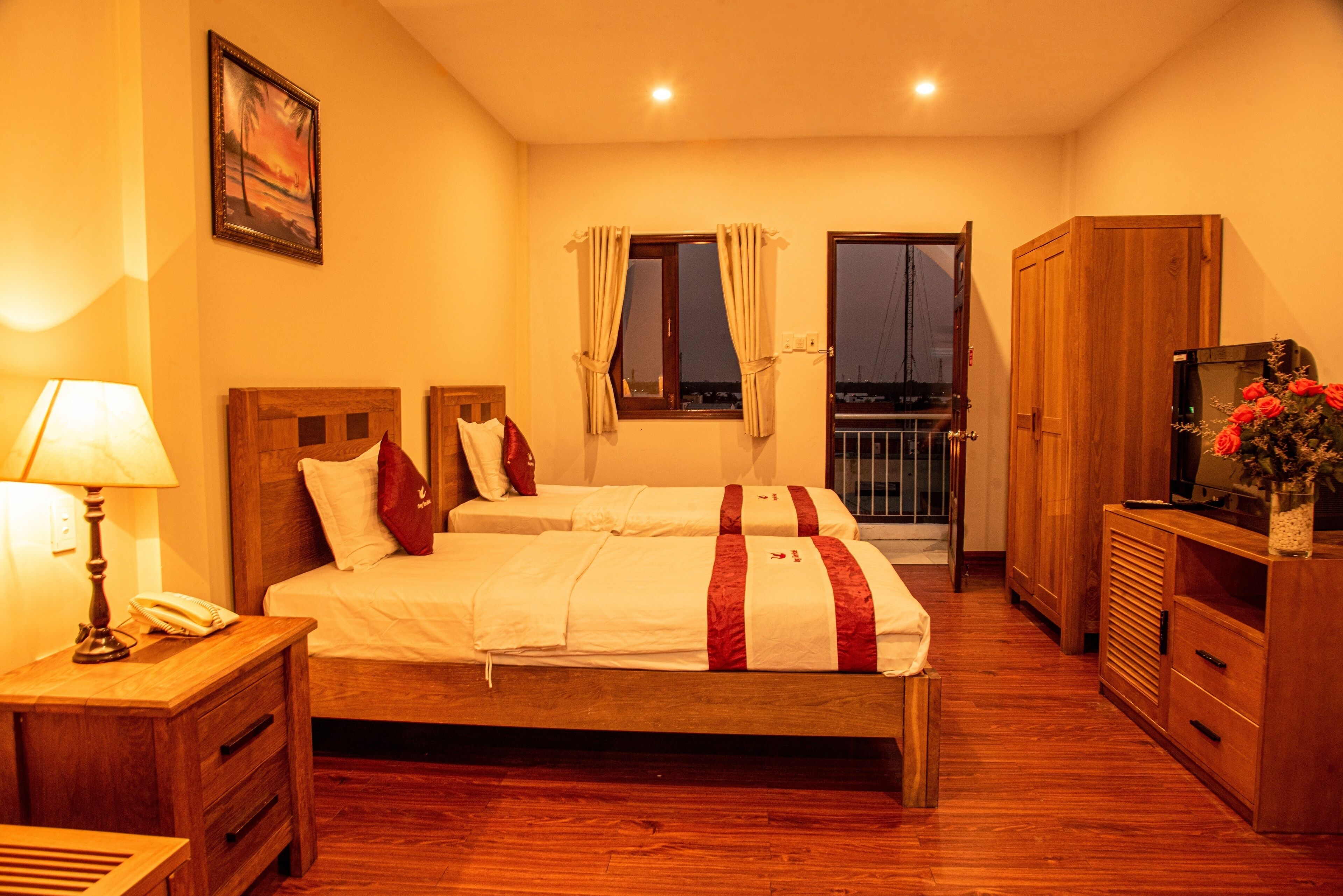 A bedroom with two beds, wooden furniture, a nightstand with a lamp, and a view of the outside through a window.