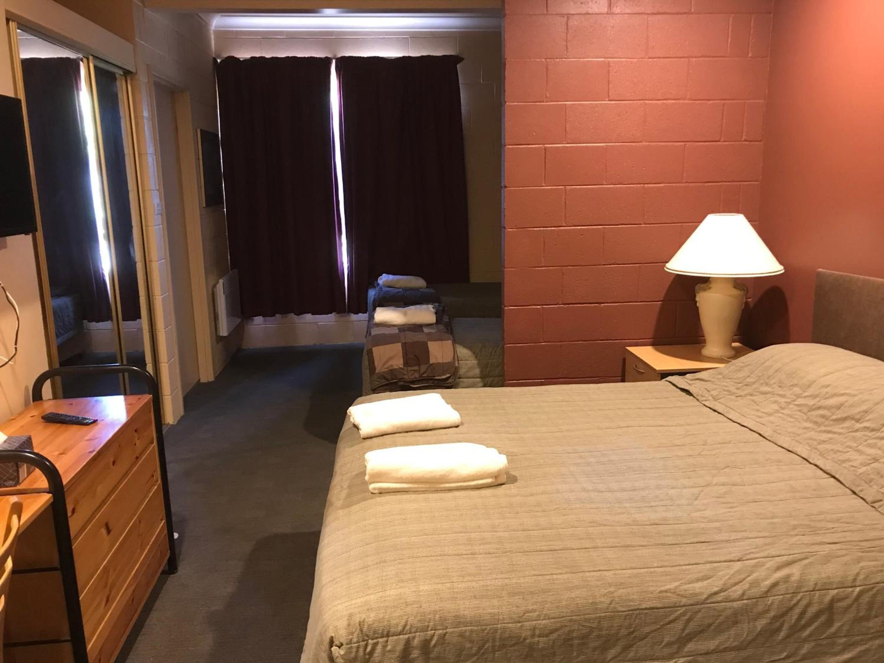 Une chambre d’hôtel avec un lit, un bureau, une chaise, une lampe et une fenêtre avec des rideaux.
