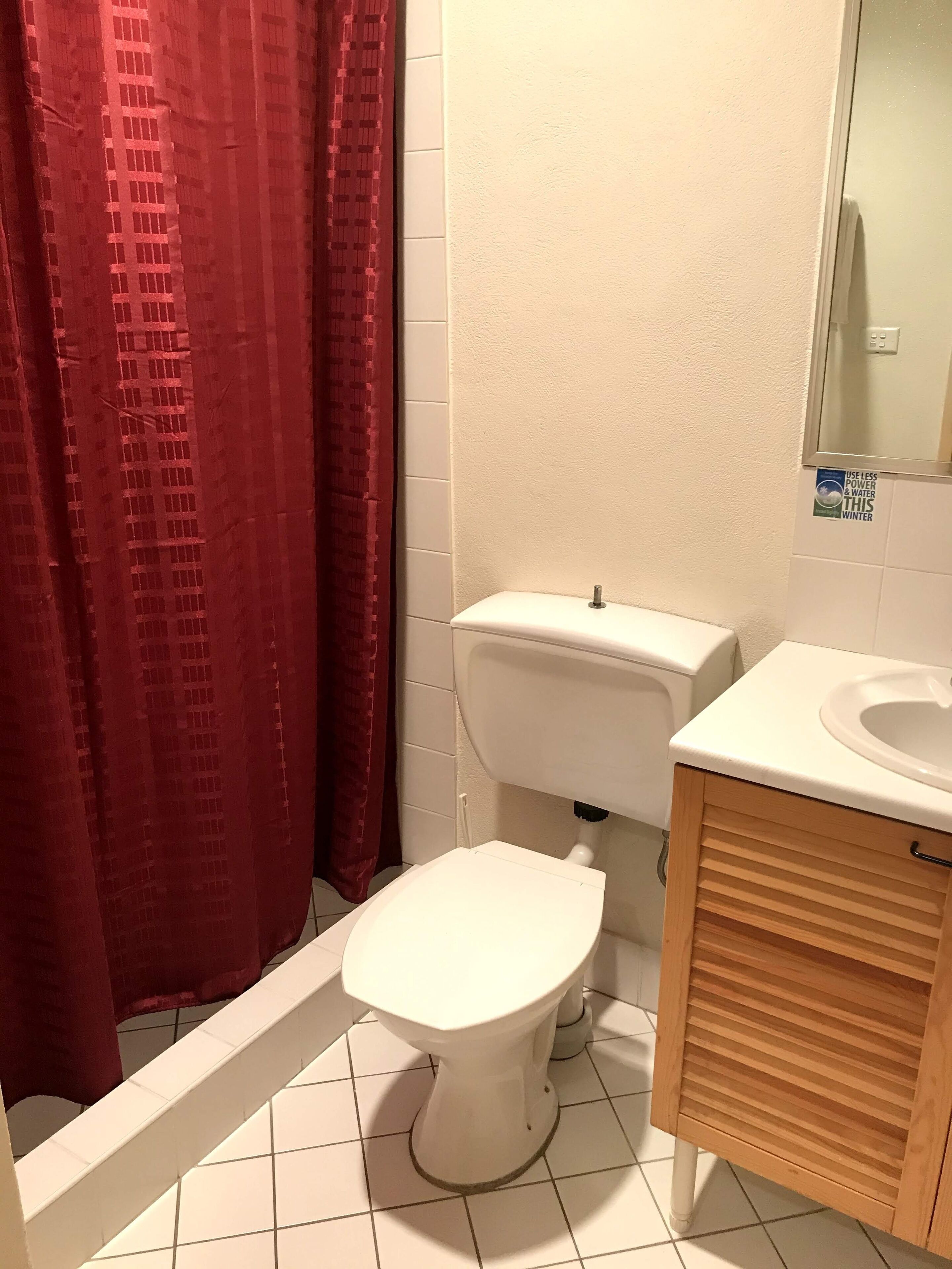 Une salle de bain avec des toilettes blanches, un meuble en bois et un rideau de douche rouge.
