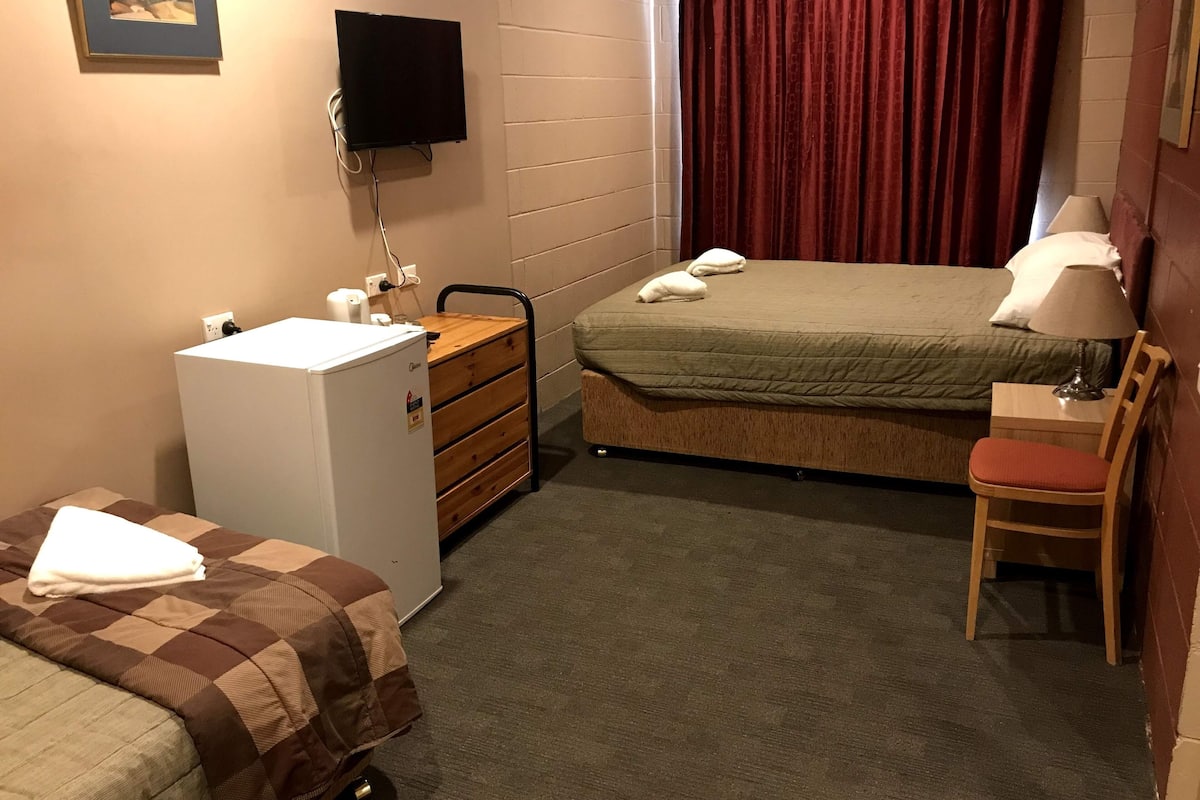A small, well-lit room with a bed, a desk, a chair, a refrigerator, and a television mounted on the wall.
