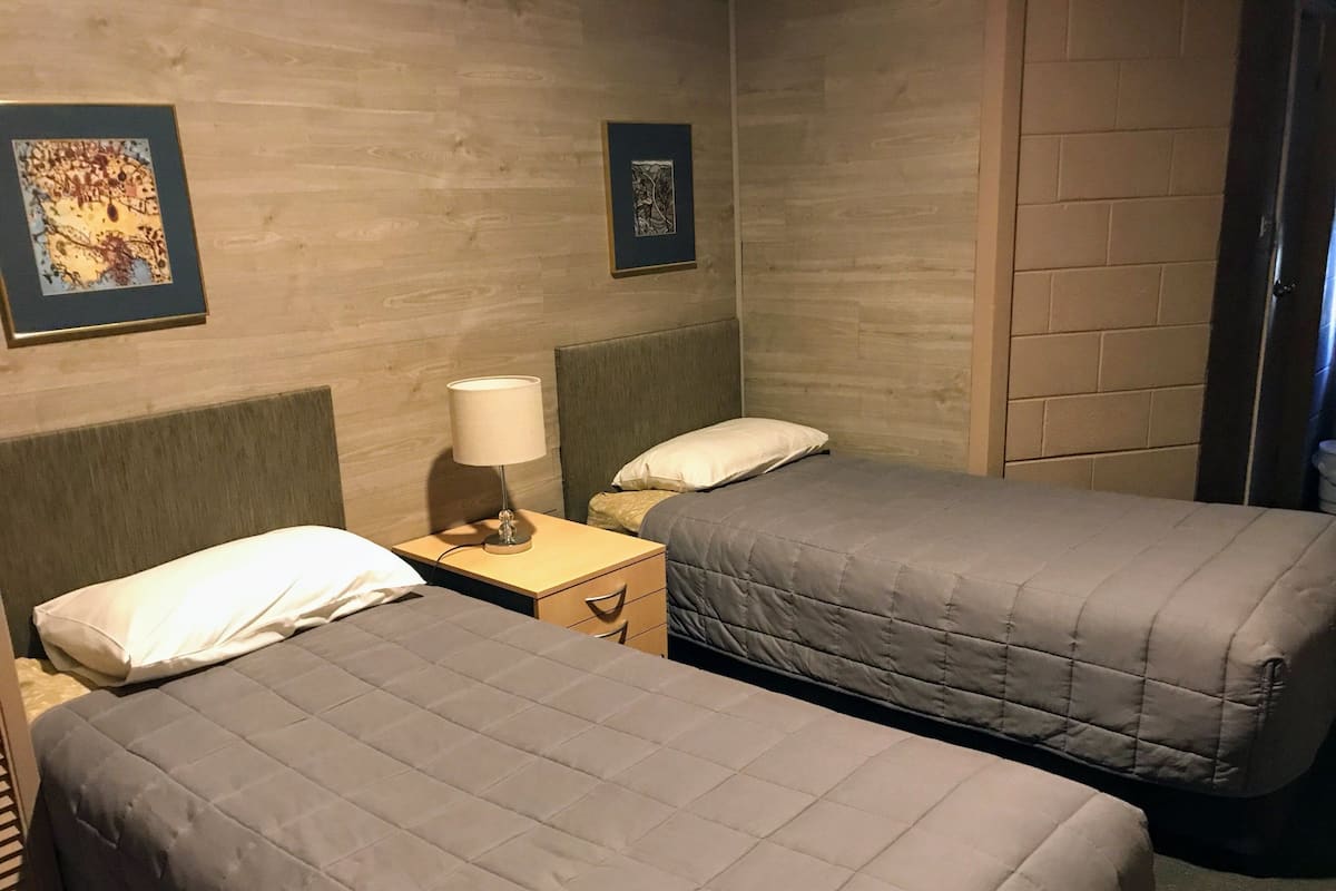 A hotel room with two single beds, a nightstand with a lamp, and two framed artworks on the wall.