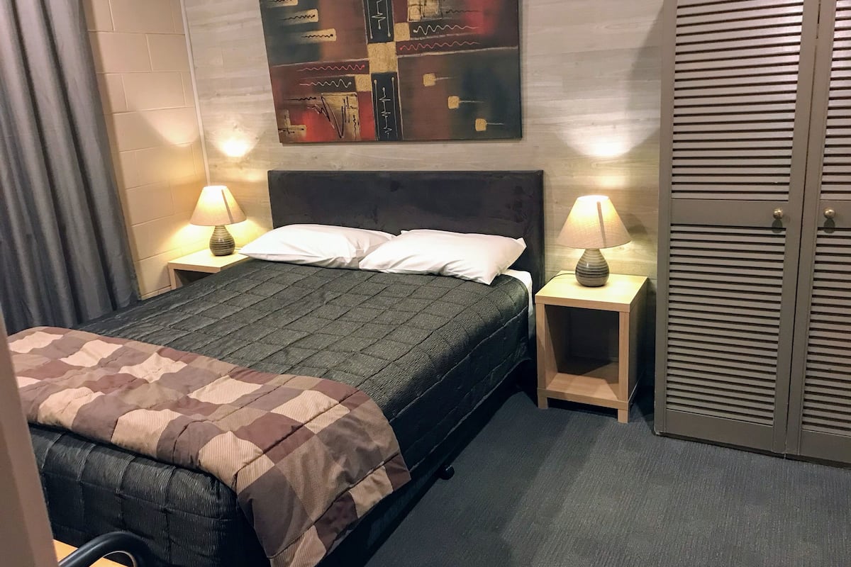A neatly made bed with a dark green quilt, two white pillows, and a brown and beige checkered throw. There are two bedside tables with lamps, a wall-mounted abstract art piece, and a wooden door with horizontal slats.