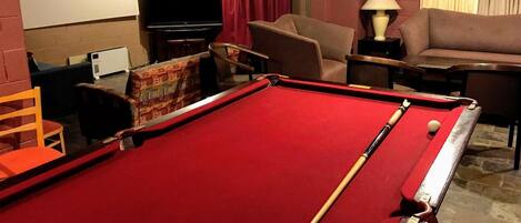Ein Billardtisch mit roter Spielunterlage, eine Poolqueue und ein Billardball. Im Hintergrund steht eine braune Couch und ein kleiner Tisch mit einer Lampe.