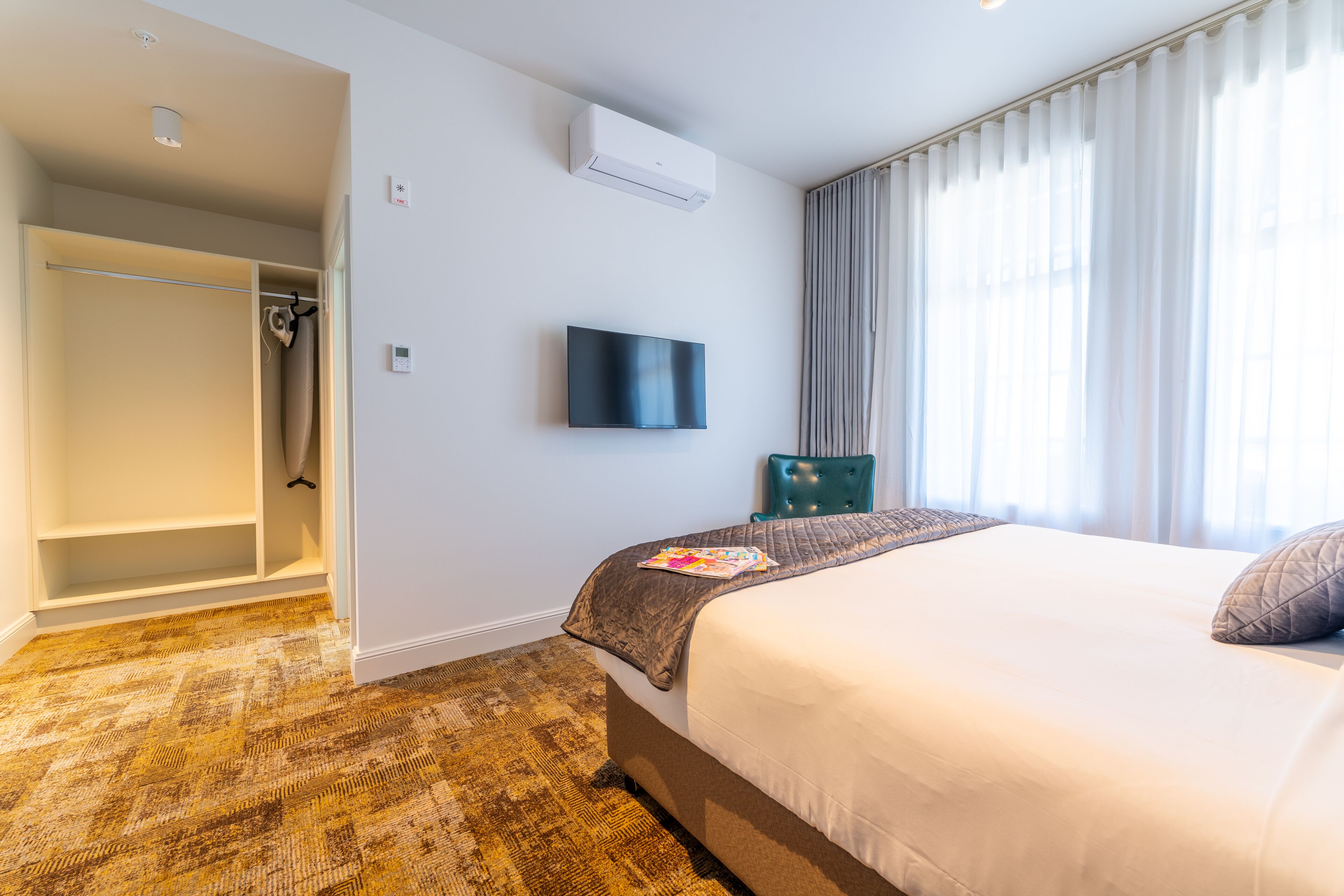Habitación de hotel con cama, televisor montado en la pared, armario y ventana con cortinas.