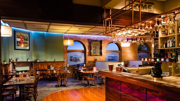 A bar area with wooden flooring, a curved bar counter, and a variety of bottles and glasses on shelves. - The Grosvenor (Timaru)