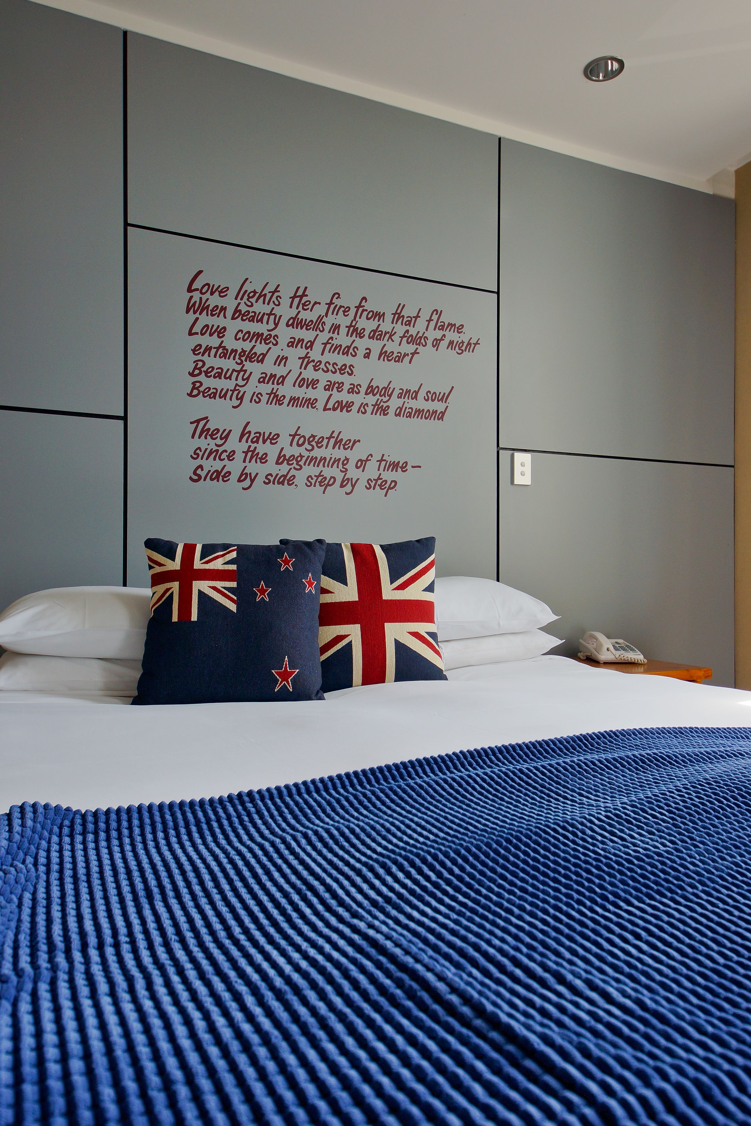A neatly made bed with white linens and a blue blanket, a pillow with a Union Jack design, and a motivational quote on the wall.