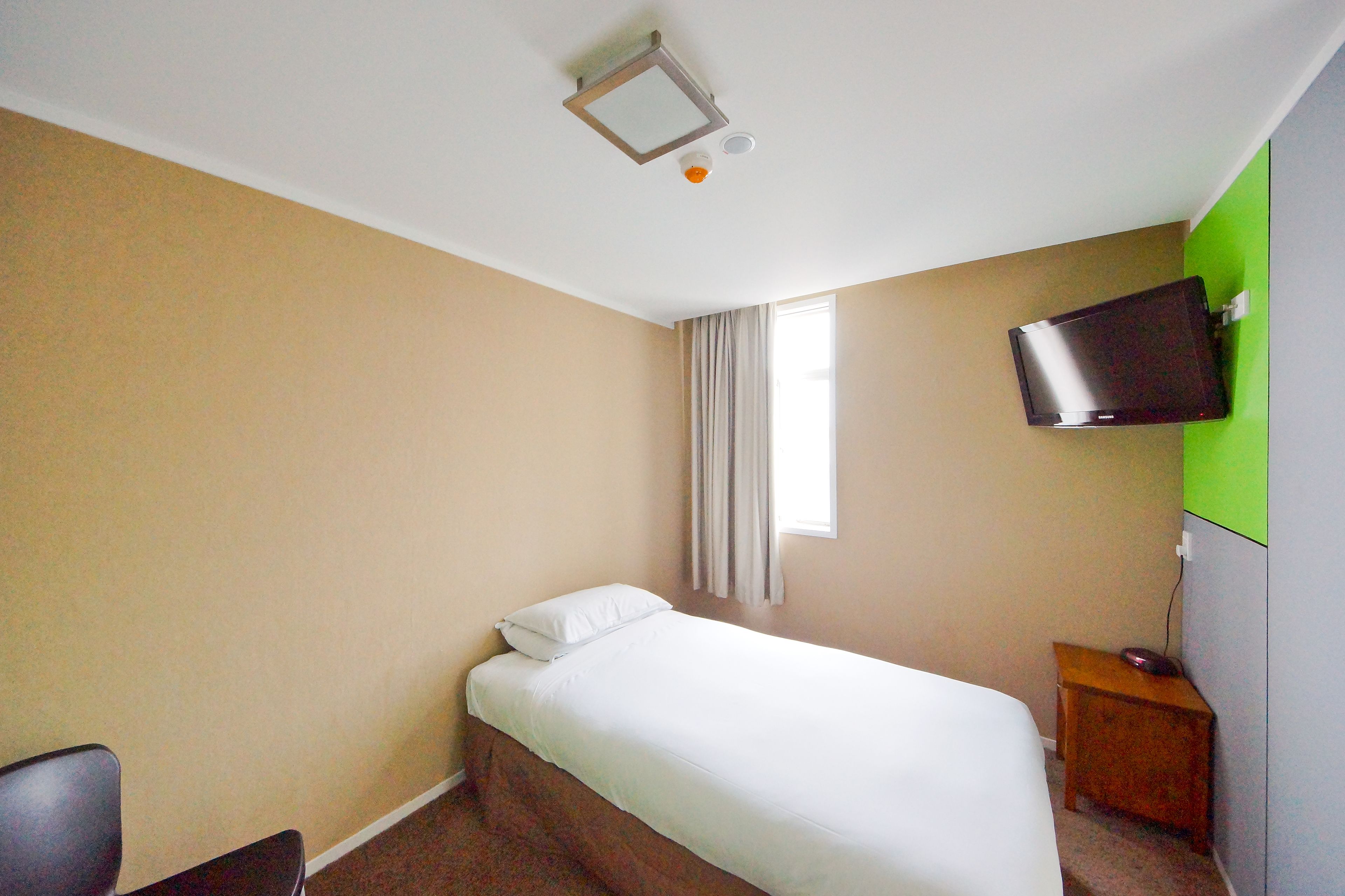 A hotel room with a bed, a television mounted on the wall, a small wooden nightstand, and a window with curtains.