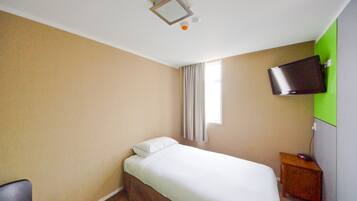 Una habitació d'hotel amb un llit, una televisió penjada a la paret, una petita tauleta de nit de fusta i una finestra amb cortines.