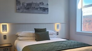 A bedroom with a bed, a nightstand, a wall-mounted light, a window, and a picture on the wall. - The Grosvenor (Timaru)