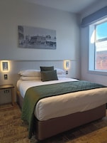 Un dormitorio con una cama, una mesita de noche, una lámpara de pared, un ventanal y un cuadro en la pared.
