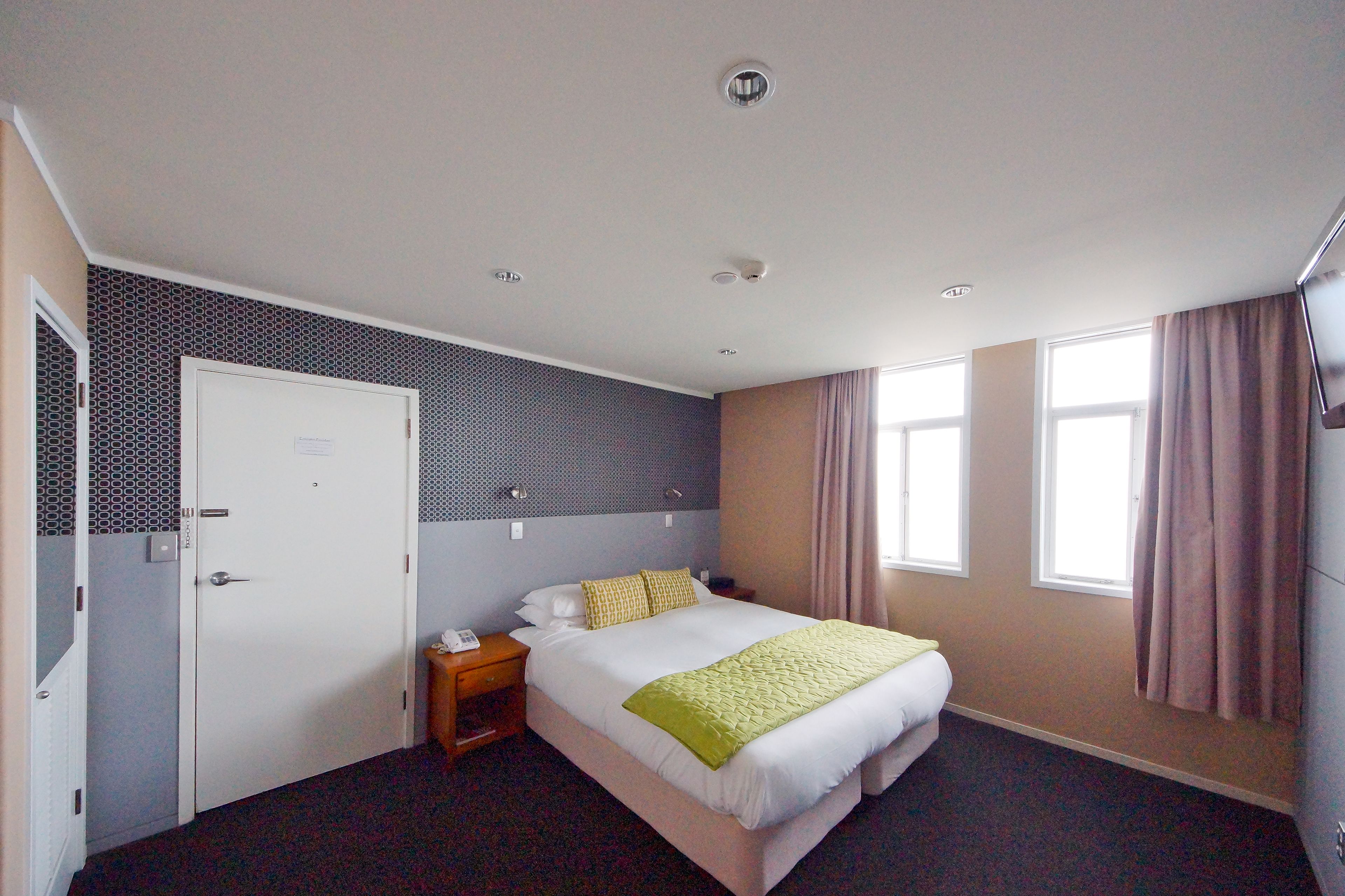 Quarto de hotel com uma cama, mesa de cabeceira, porta e duas janelas com cortinas.