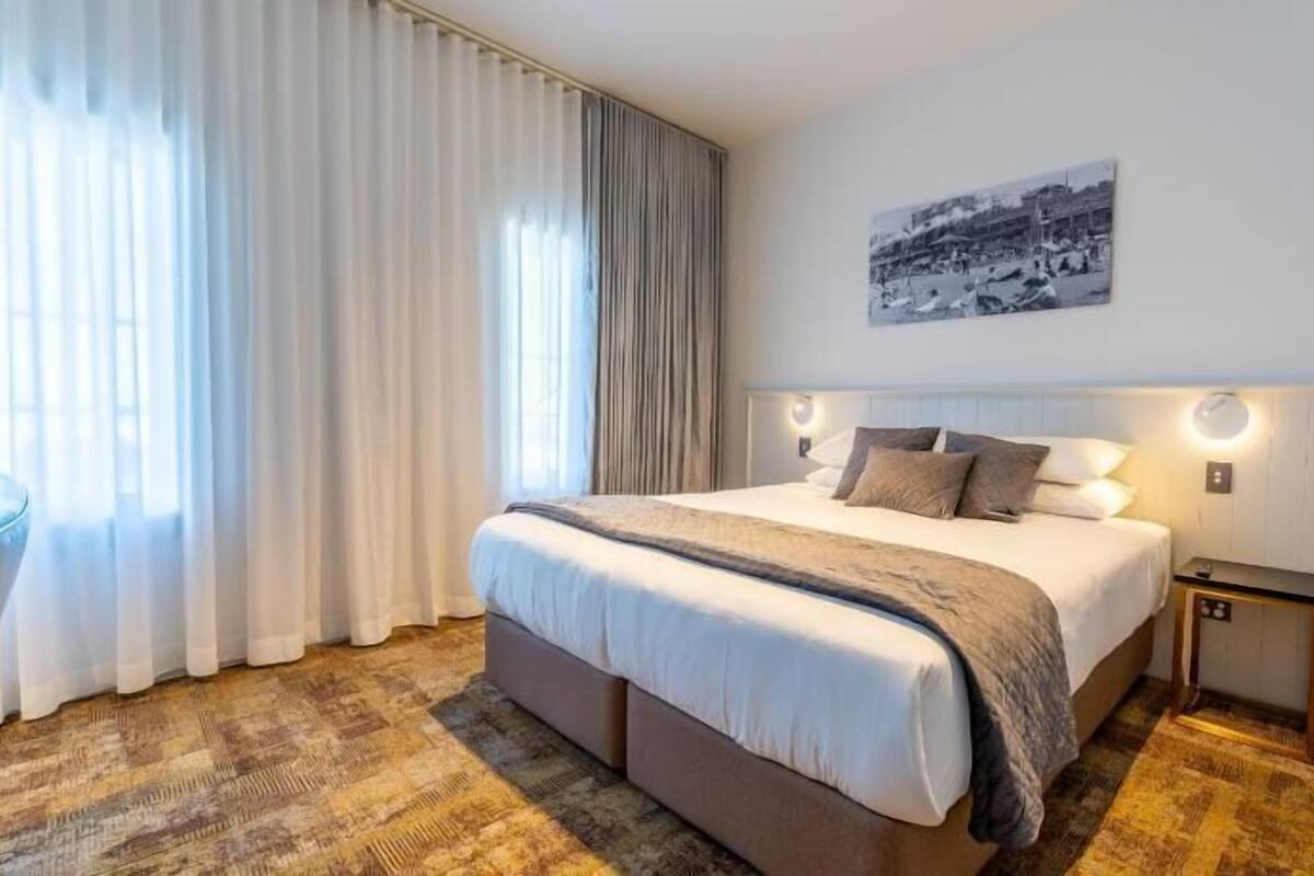 A hotel room with a large bed, a side table, a nightstand, a wall-mounted light, and a window with curtains.