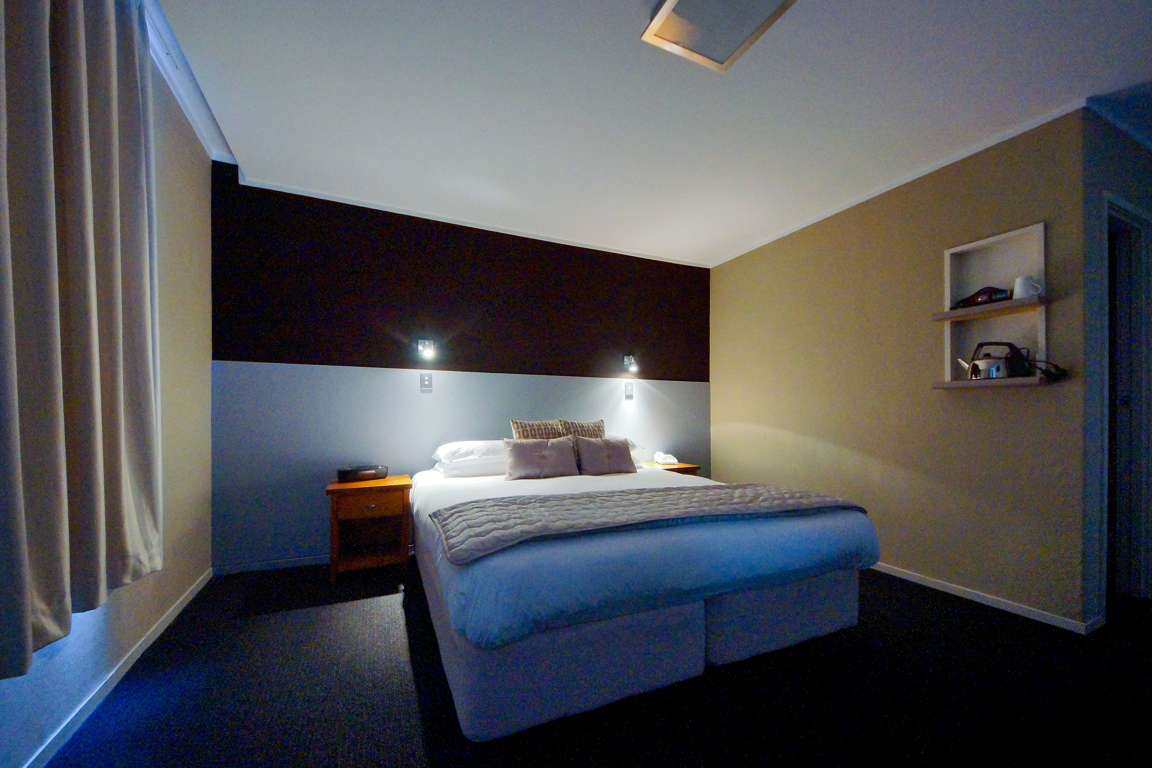 A hotel room with a bed, bedside tables, a nightstand, a shelf with toiletries, and a window with curtains.