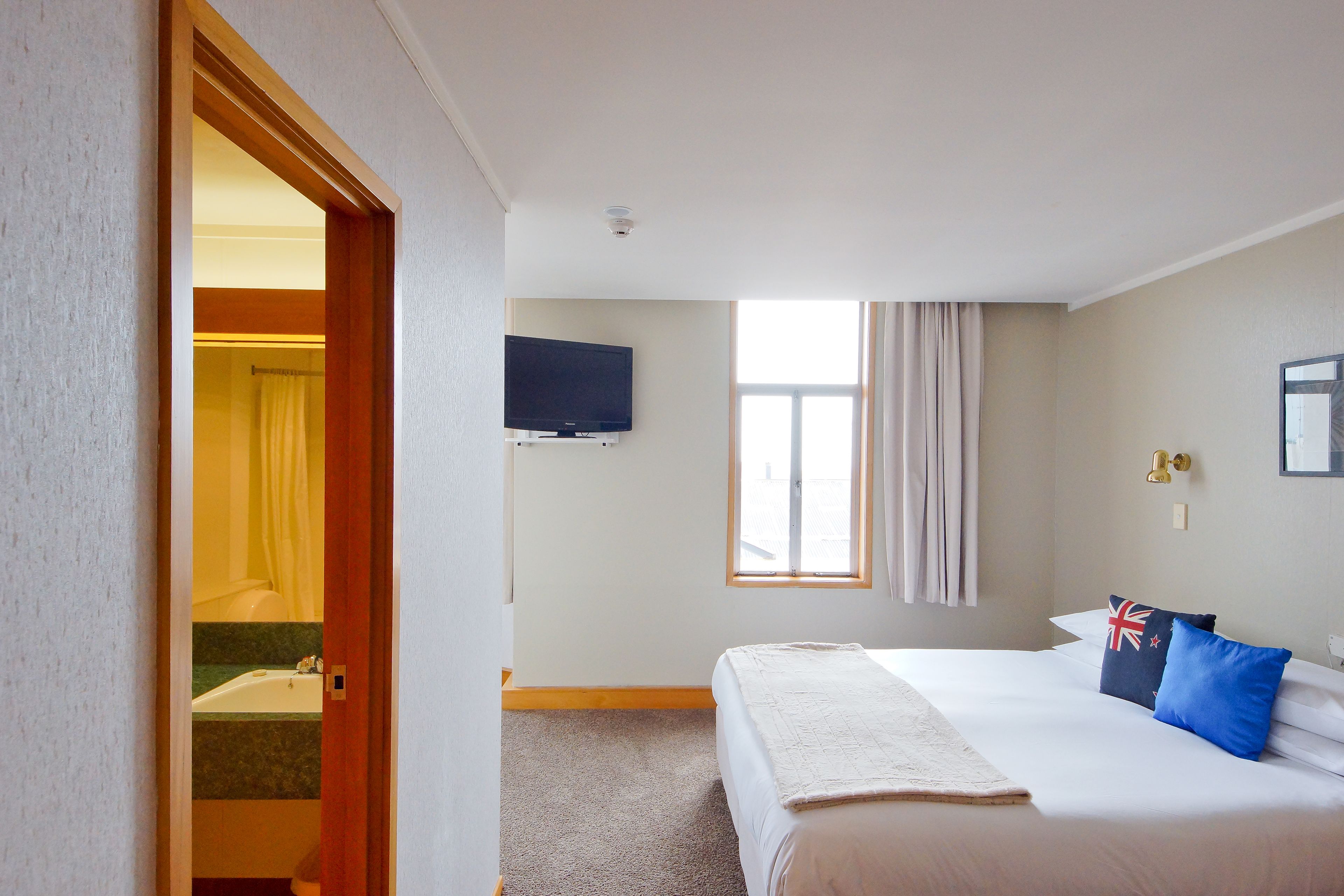 Habitación de hotel con cama, televisión en la pared, ventana con cortinas y baño visible a través de una puerta abierta.