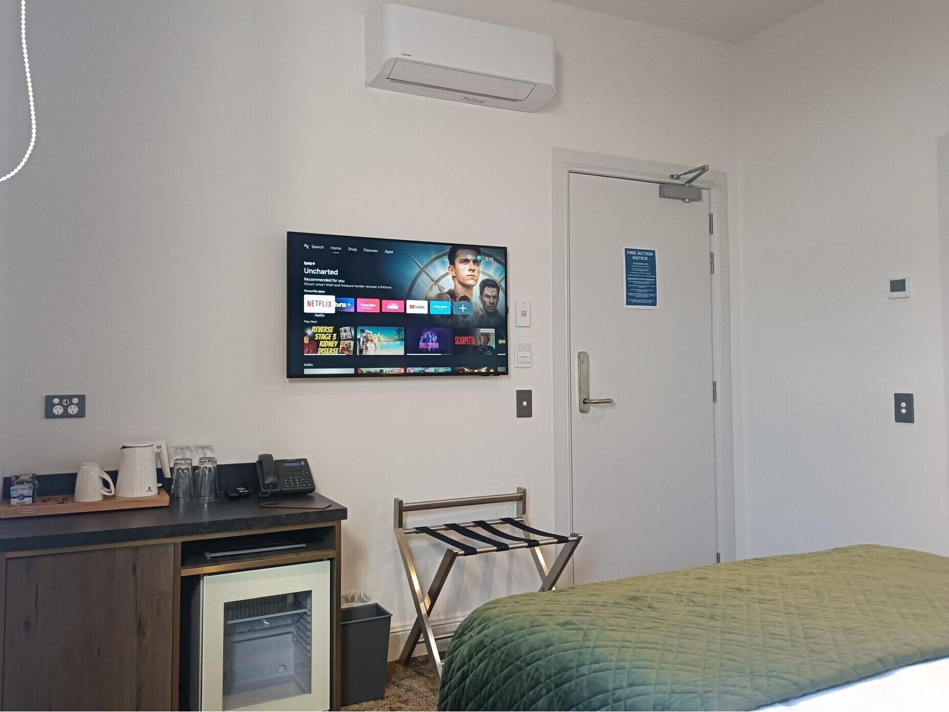 Une chambre d’hôtel dotée d’un téléviseur à écran plat fixé au mur, d’un bureau avec une machine à café, d’un mini-réfrigérateur, d’un étendoir et d’un lit recouvert d’une couette verte.