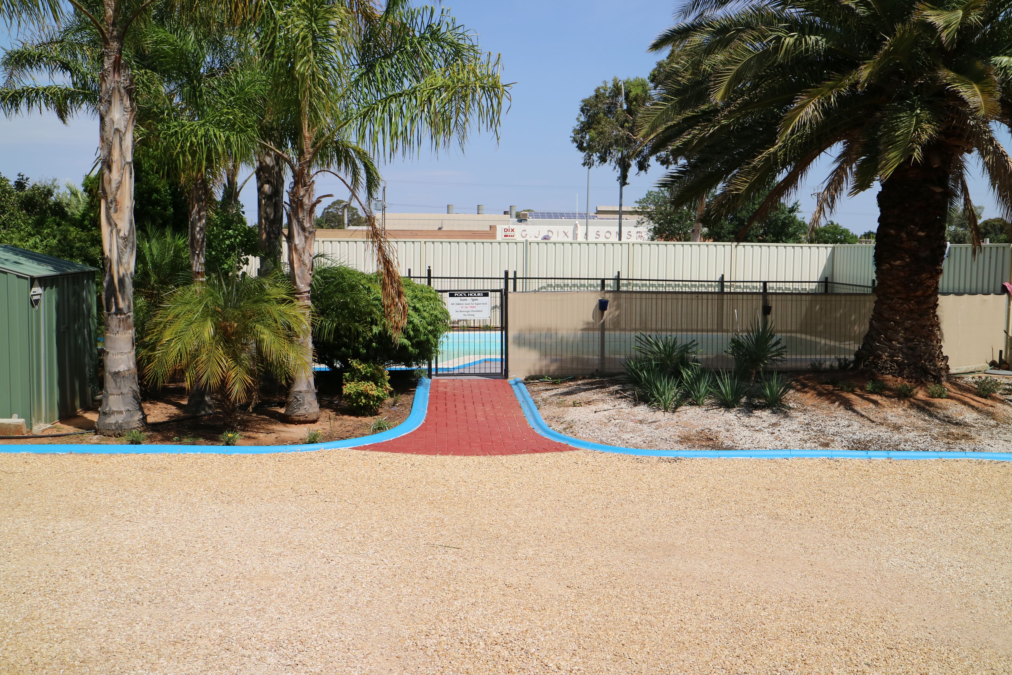 A gravel path leading to a gated area with palm trees and a building in the background.