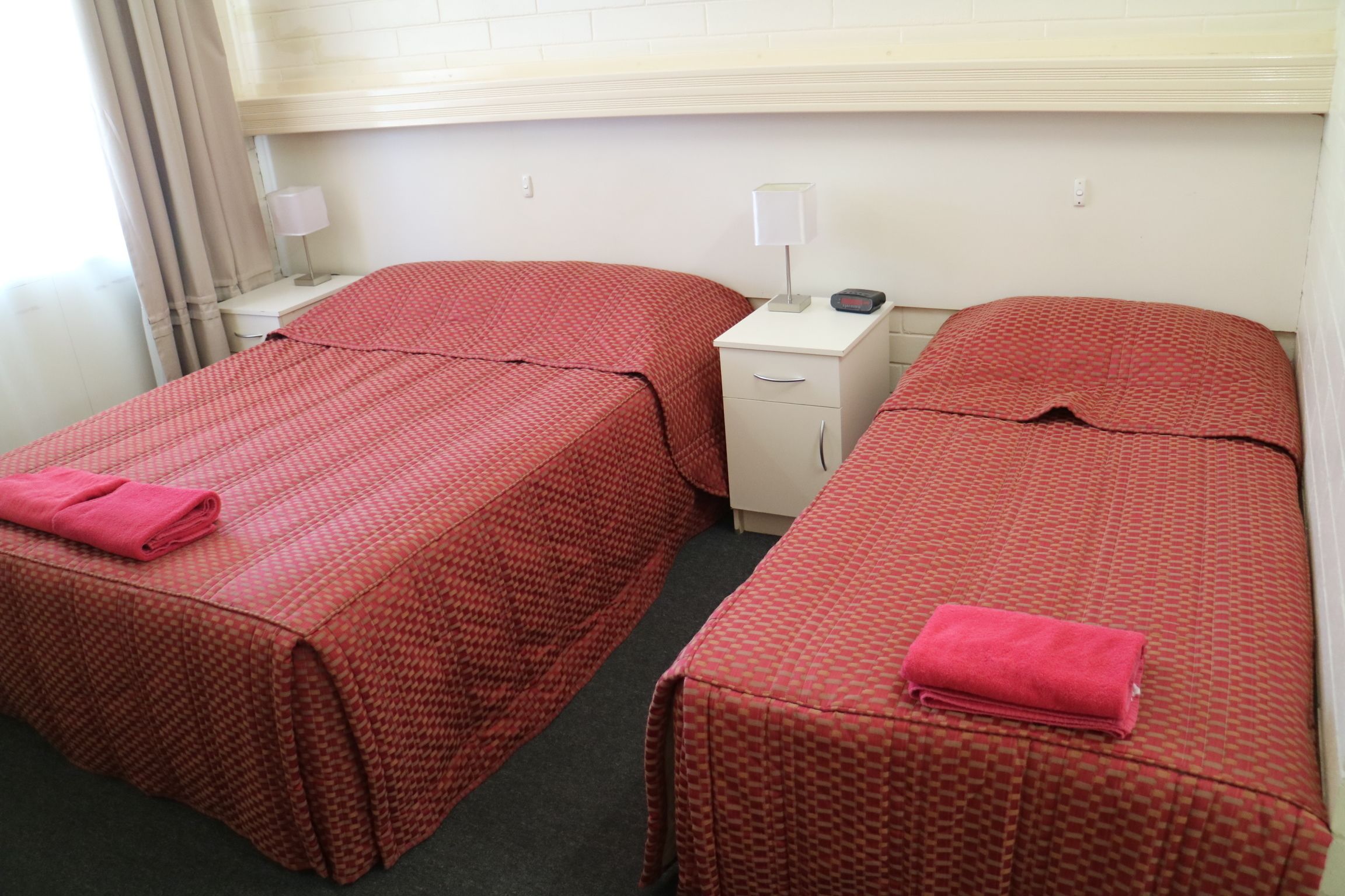 Une chambre d’hôtel avec deux lits, chacun recouvert d’une couvre-lit à motifs rouges et agrémenté d’une serviette rose. Il y a deux tables de chevet avec des lampes et une petite horloge. La chambre présente un aménagement simple et épuré, avec des murs blancs et une fenêtre avec des rideaux.