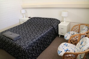 A bedroom with a black bedspread, two bedside tables with lamps, a wicker chair with a butterfly pattern, and a brown carpet.