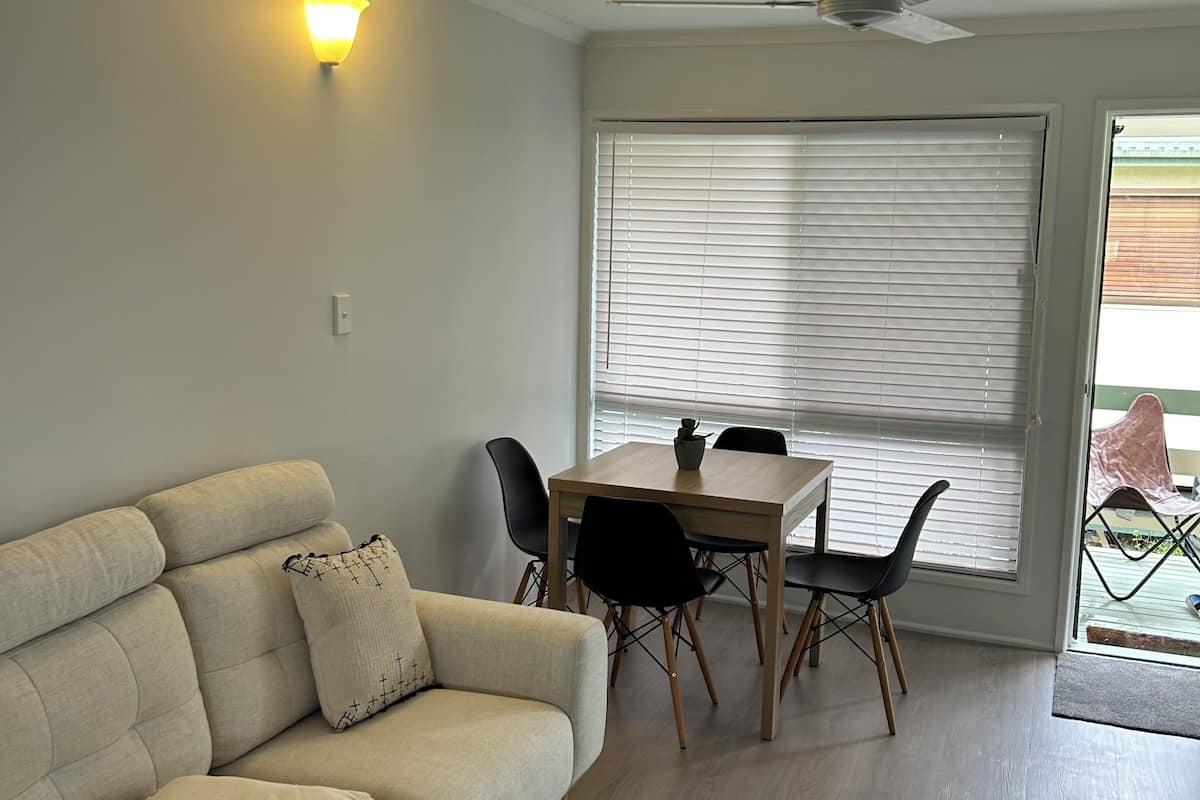 Uma sala de estar com sofá, mesa de jantar com cadeiras, ventilador de teto e janela com persianas.