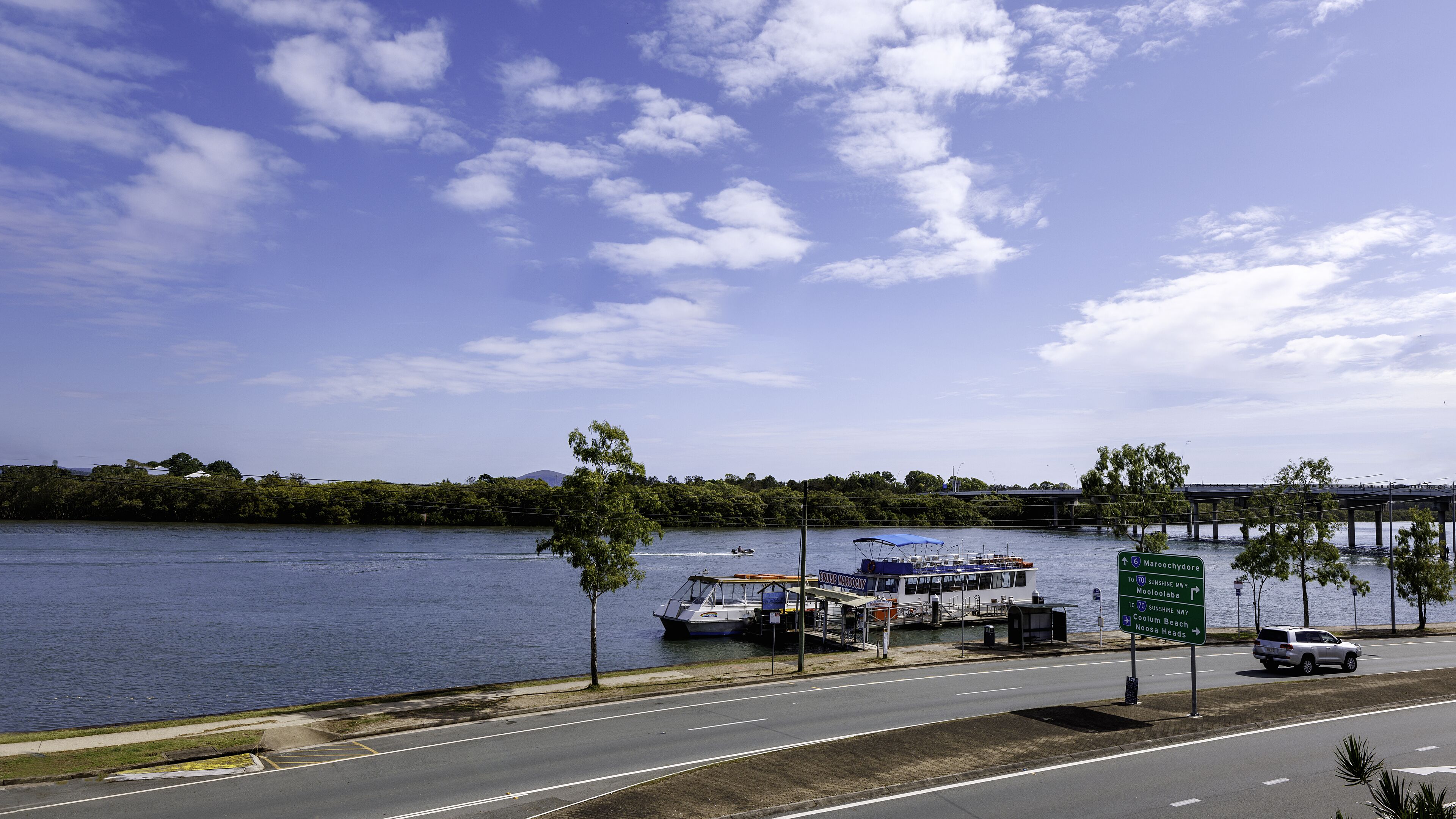 Une route longeant le bord de l’eau, avec un pont en arrière-plan, un bateau amarré dans une marina et un panneau de signalisation indiquant les directions vers différents lieux.