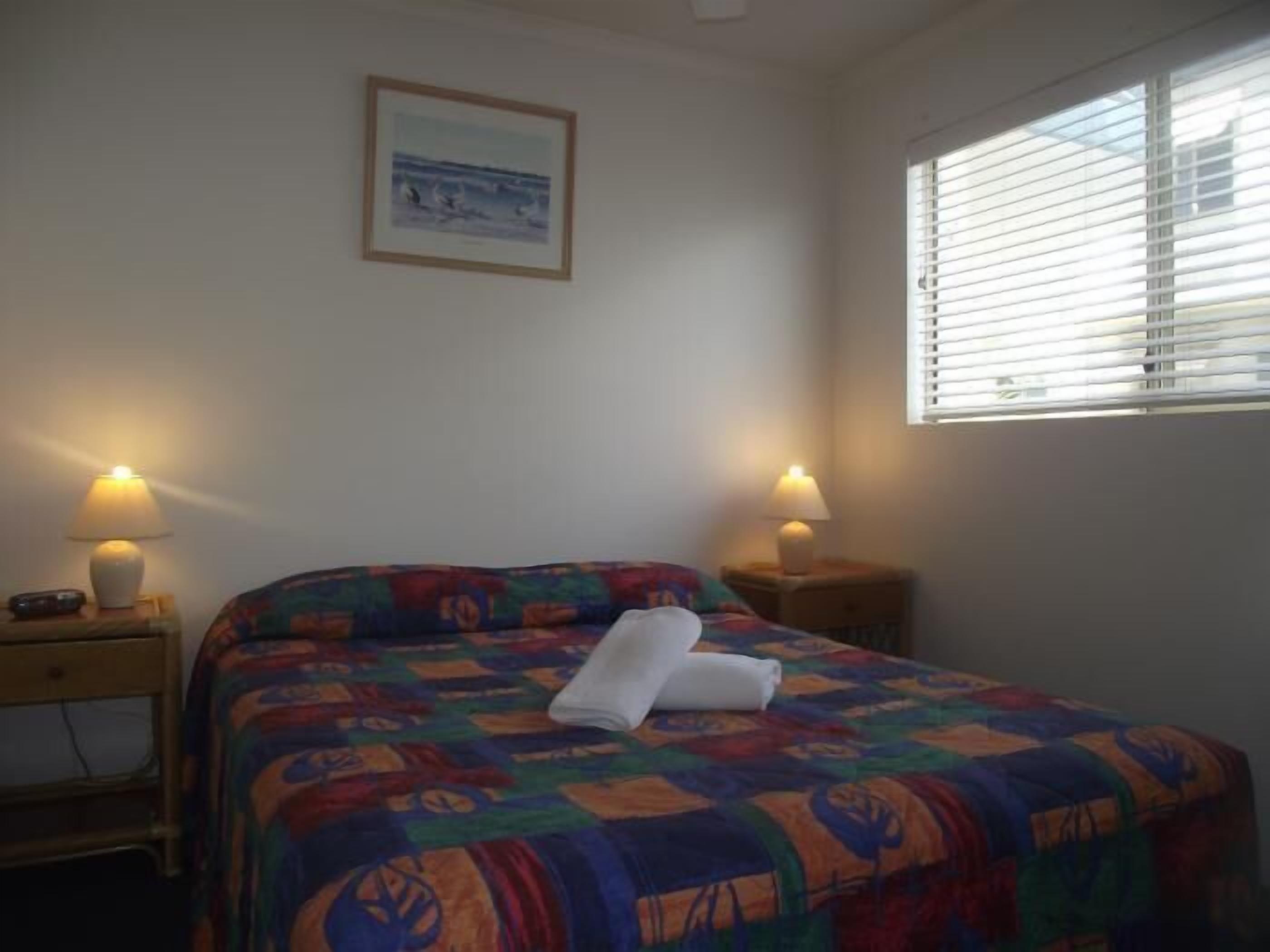 A bedroom with a colorful bedspread, two bedside lamps, a framed picture on the wall, and a window with blinds.