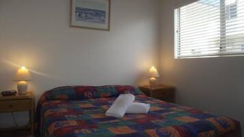 Un dormitorio con una colcha de colores, dos lámparas de noche, un cuadro enmarcado en la pared y una ventana con persianas.
