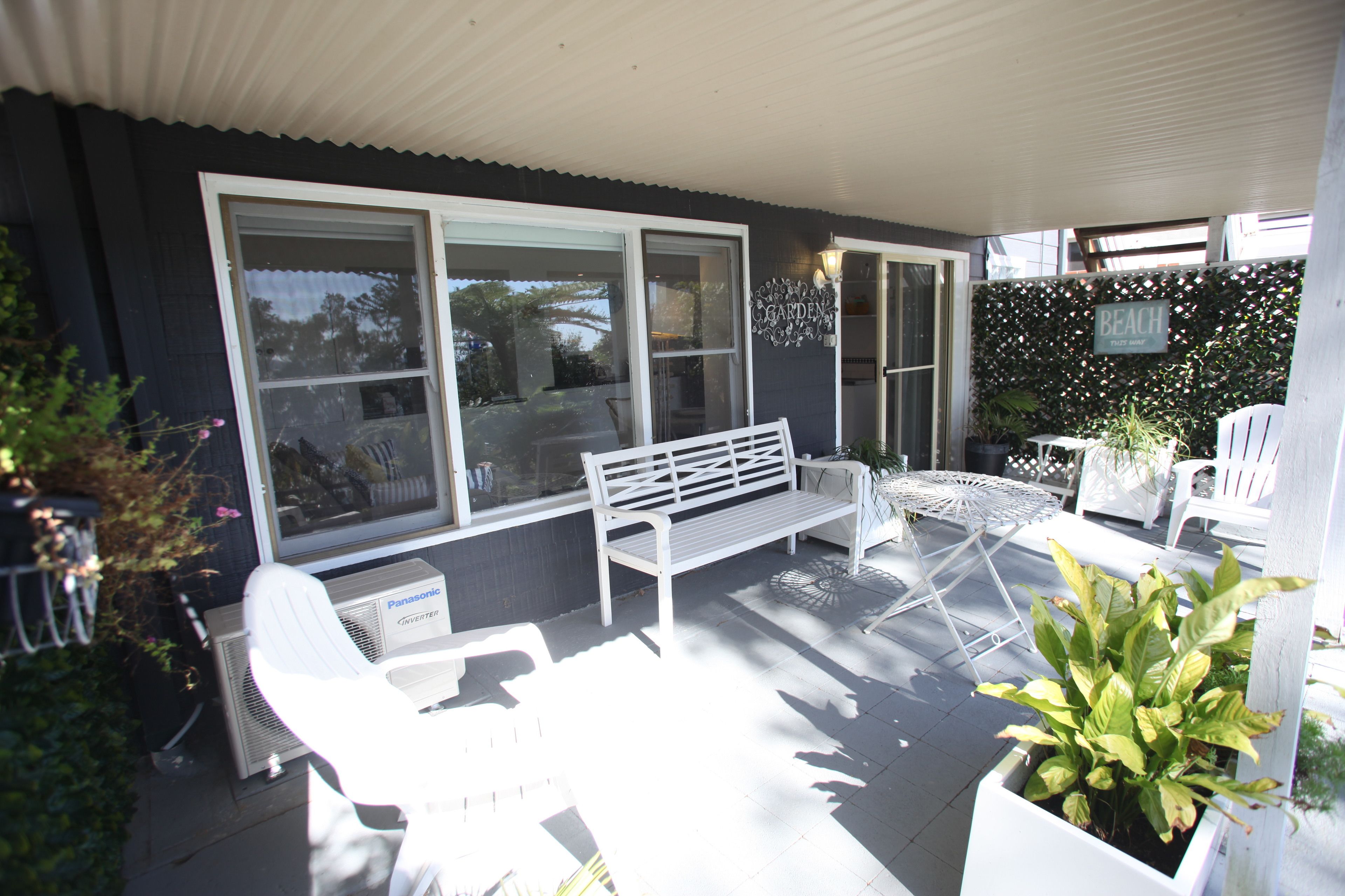 A covered patio with white furniture, including a bench and chairs, and a potted plant.