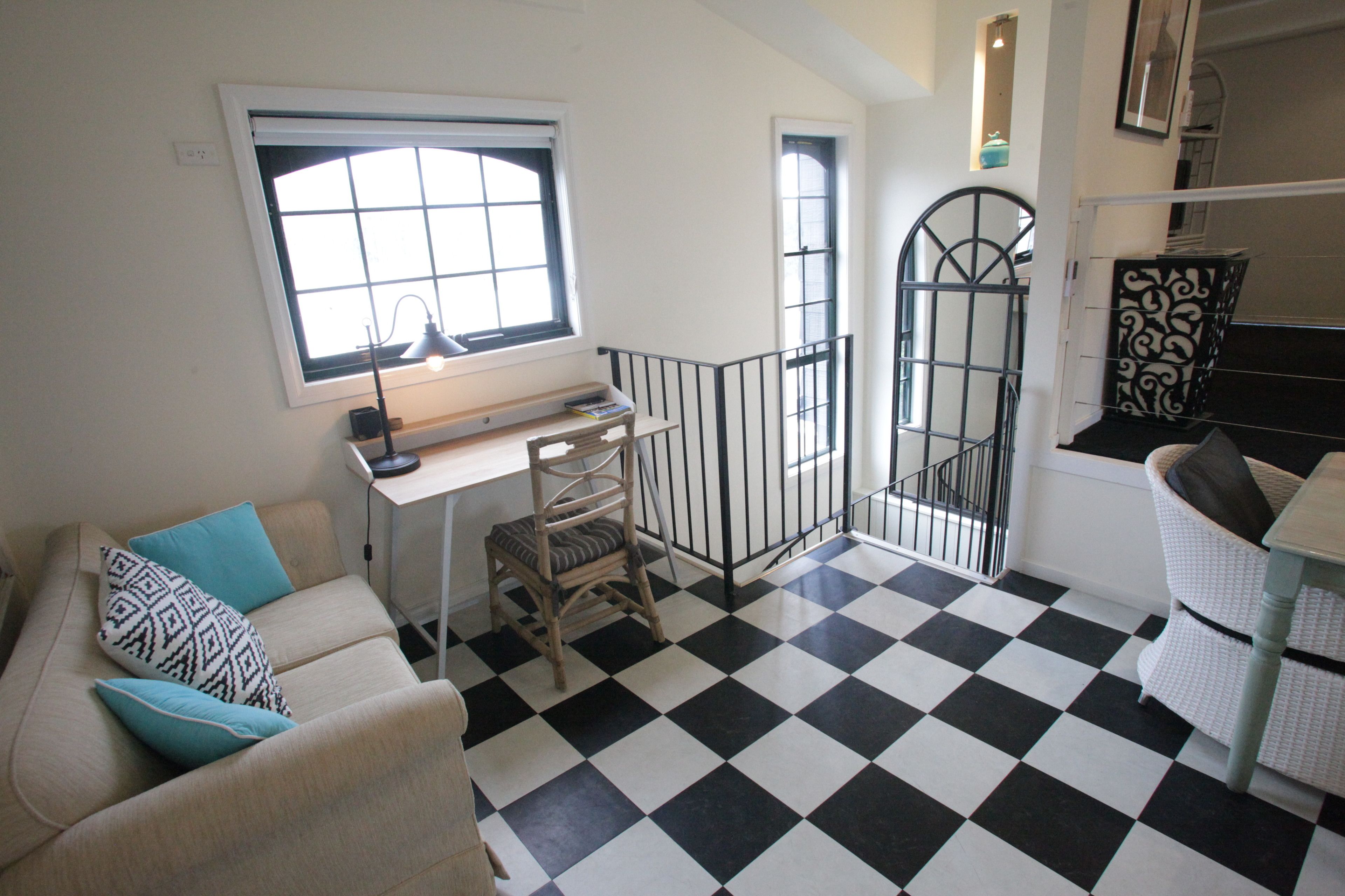 A room with a checkered floor, a desk with a lamp, a chair, a sofa with patterned cushions, and a staircase with a decorative railing.