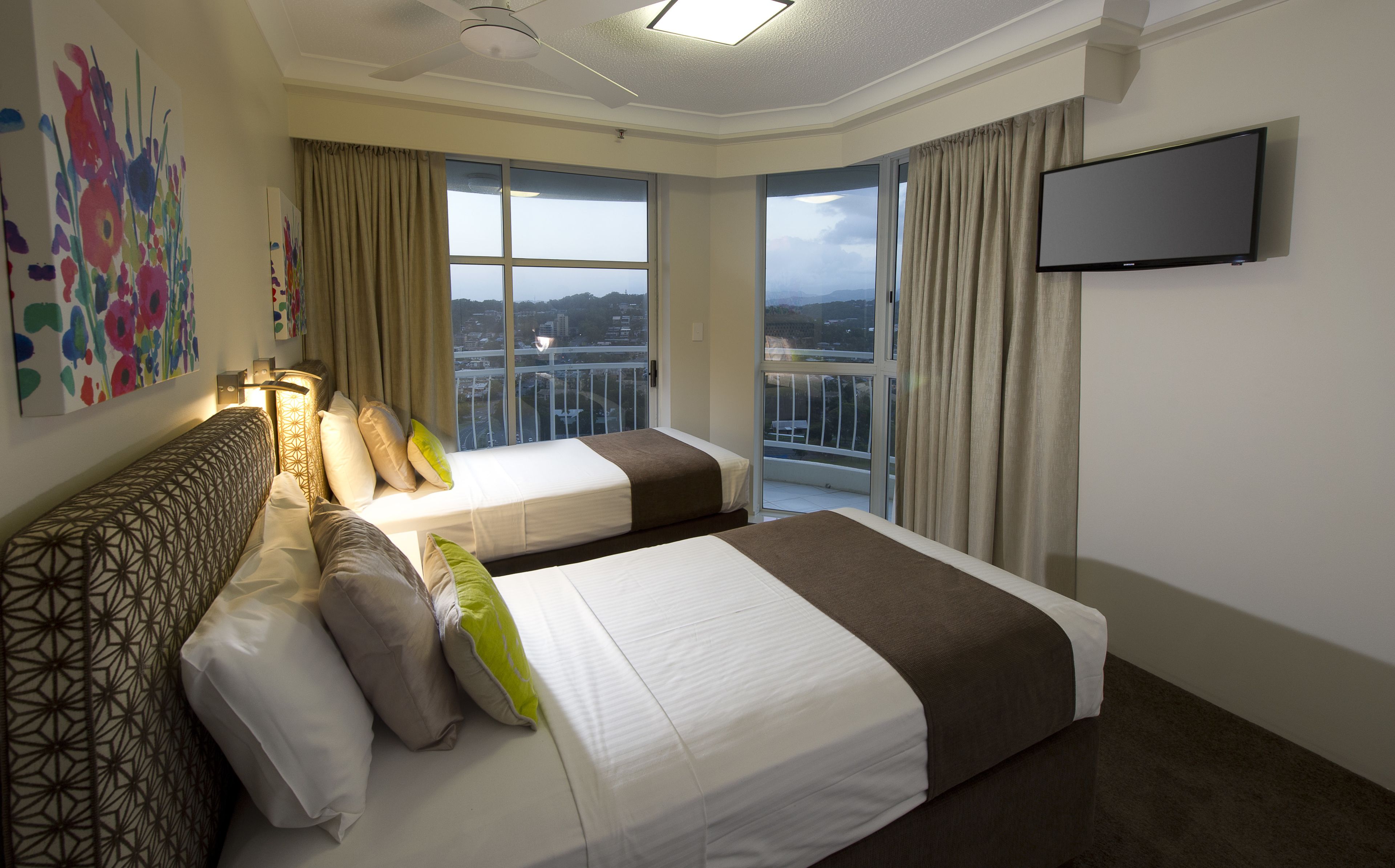 A hotel room with two beds, a large window with a city view, a flat-screen TV, and a ceiling fan.