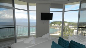 A hotel room with a large bed, white bedding, and a view of the ocean and cityscape through large windows. - 2nd Avenue Beachside Apartments (Burleigh Heads)