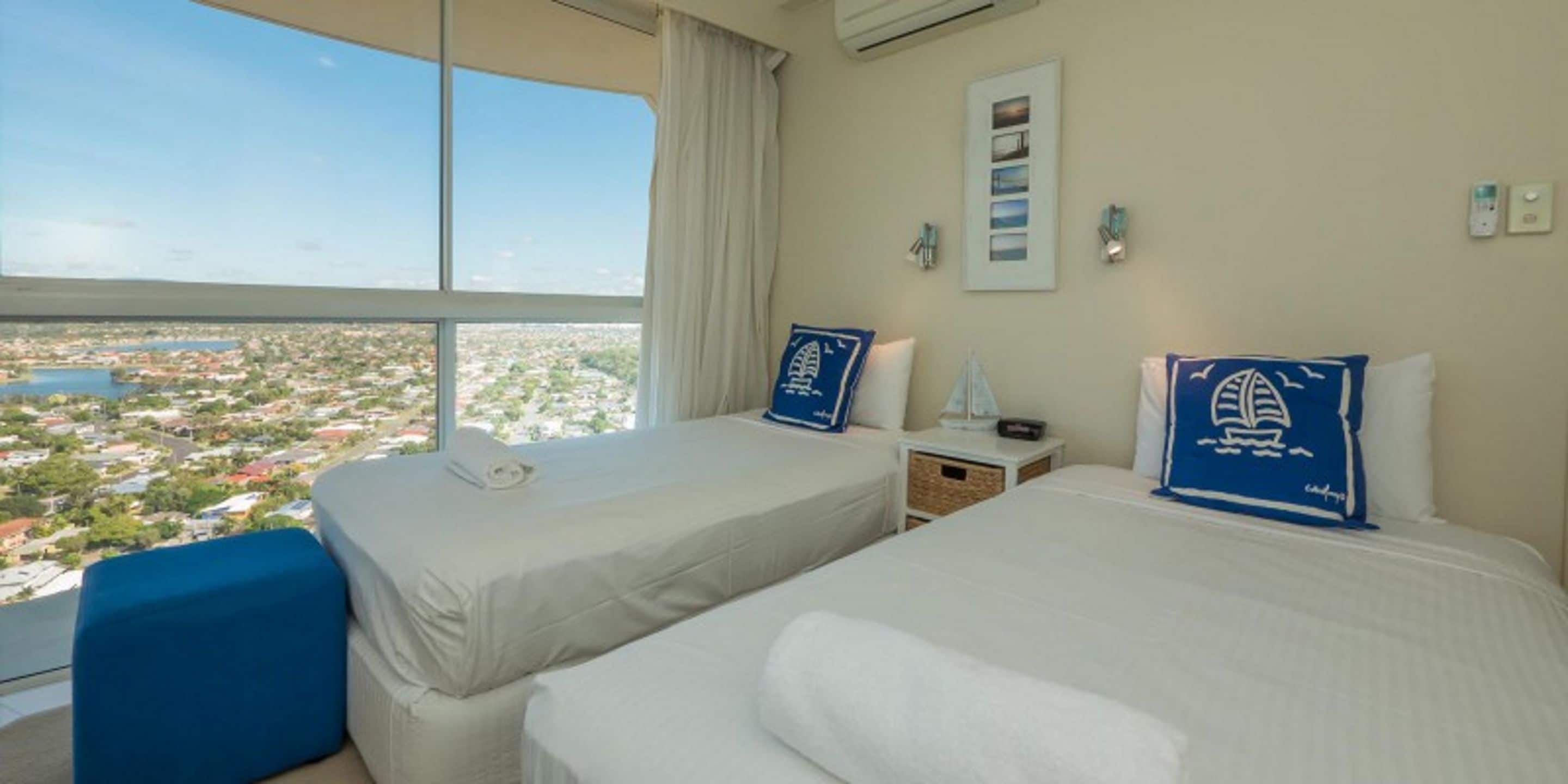 A hotel room with two beds, a large window with a city view, and a small table with a lamp.