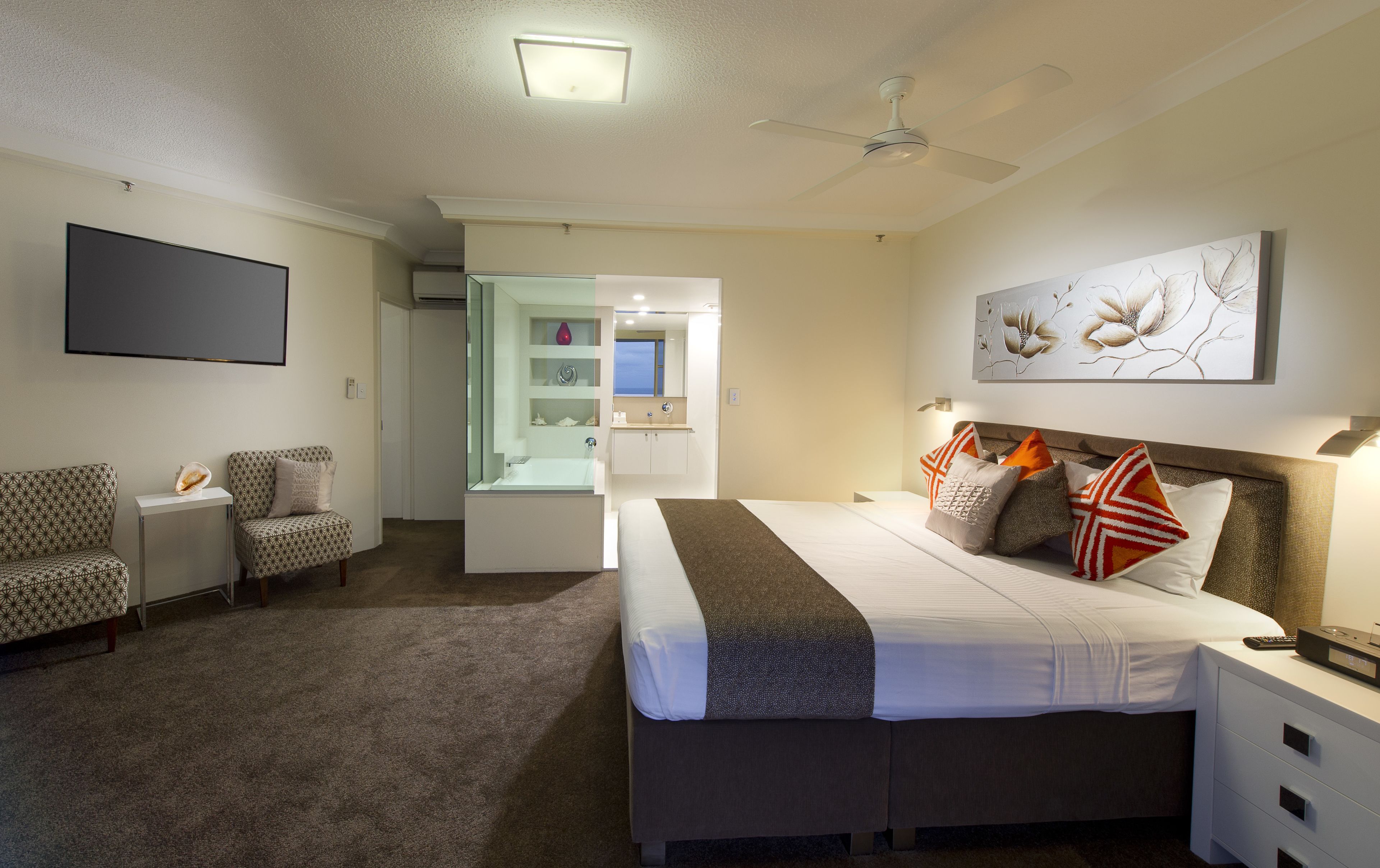 A hotel room with a bed, two armchairs, a small seating area with a glass partition, a flat-screen TV, and a ceiling fan.