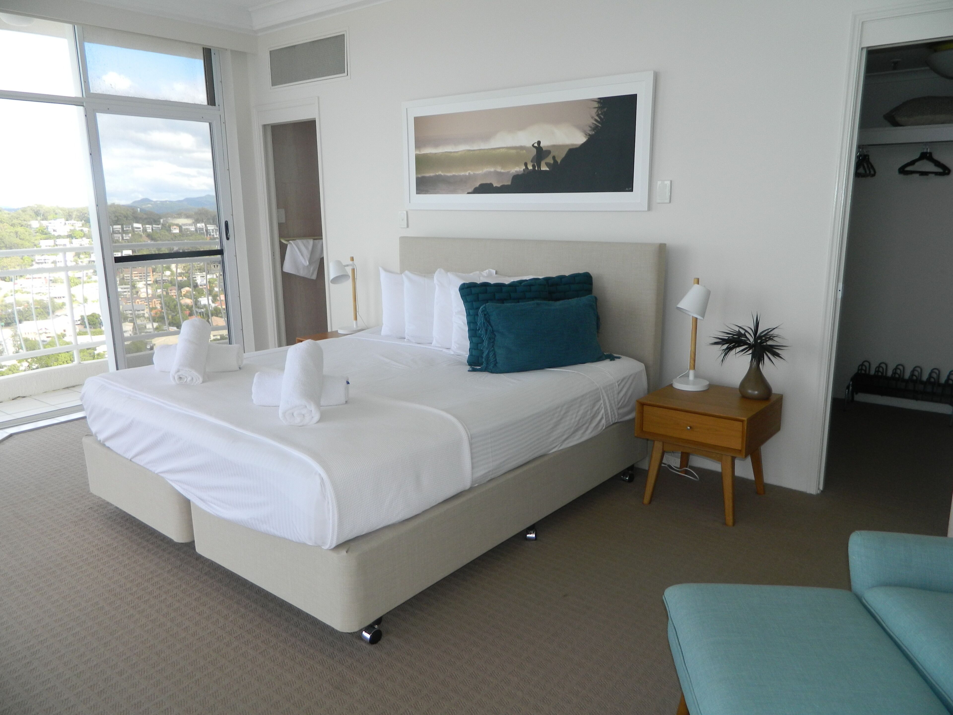 A hotel room with a large bed, a nightstand, a lamp, a painting, and a view of the city.