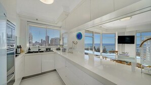 A modern kitchen with white cabinets, a built-in oven, and a large window overlooking a cityscape. - 2nd Avenue Beachside Apartments (Burleigh Heads)