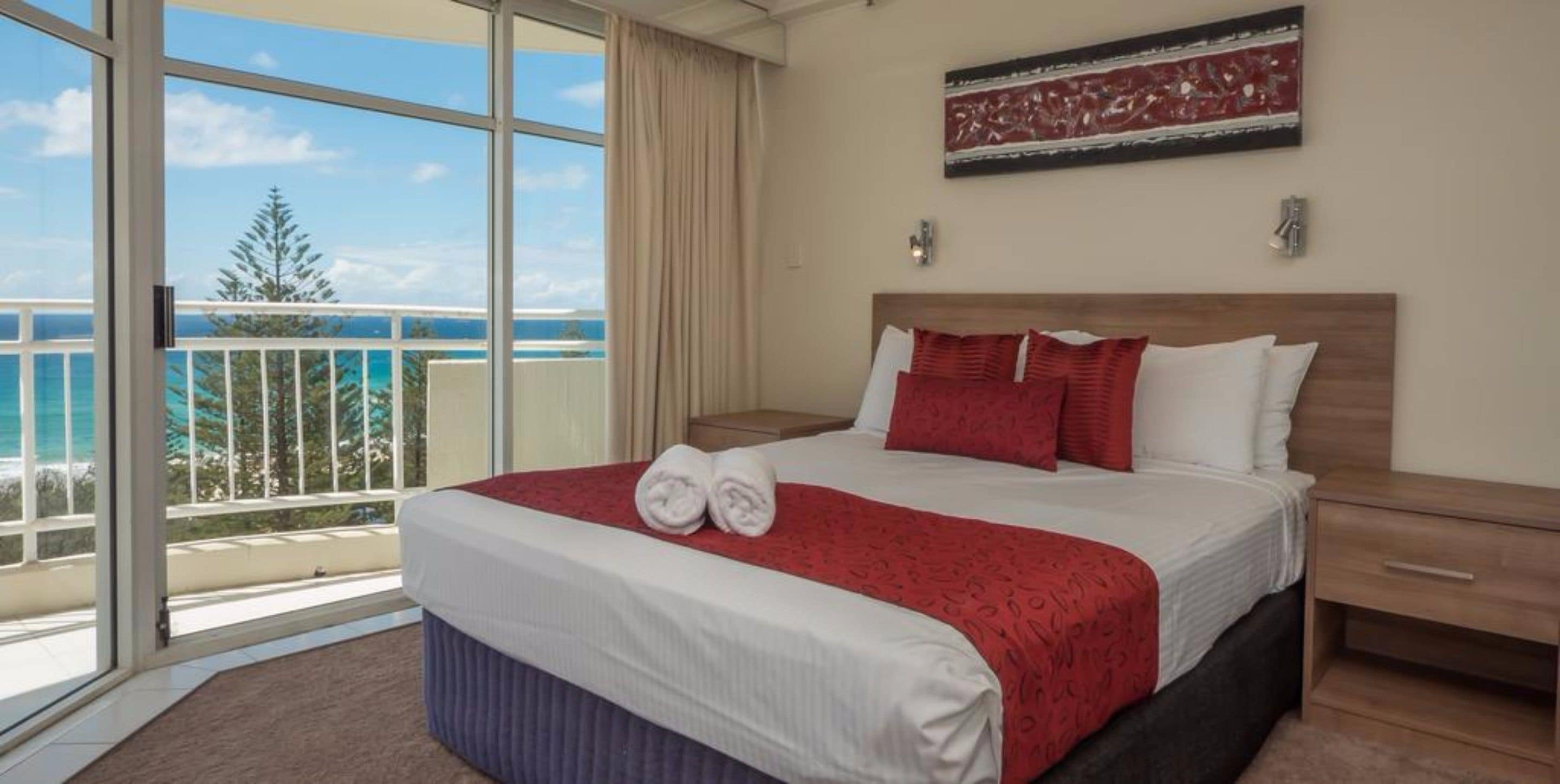 A hotel room with a large bed, a nightstand, a red and white bedspread, and a view of the ocean through a balcony.