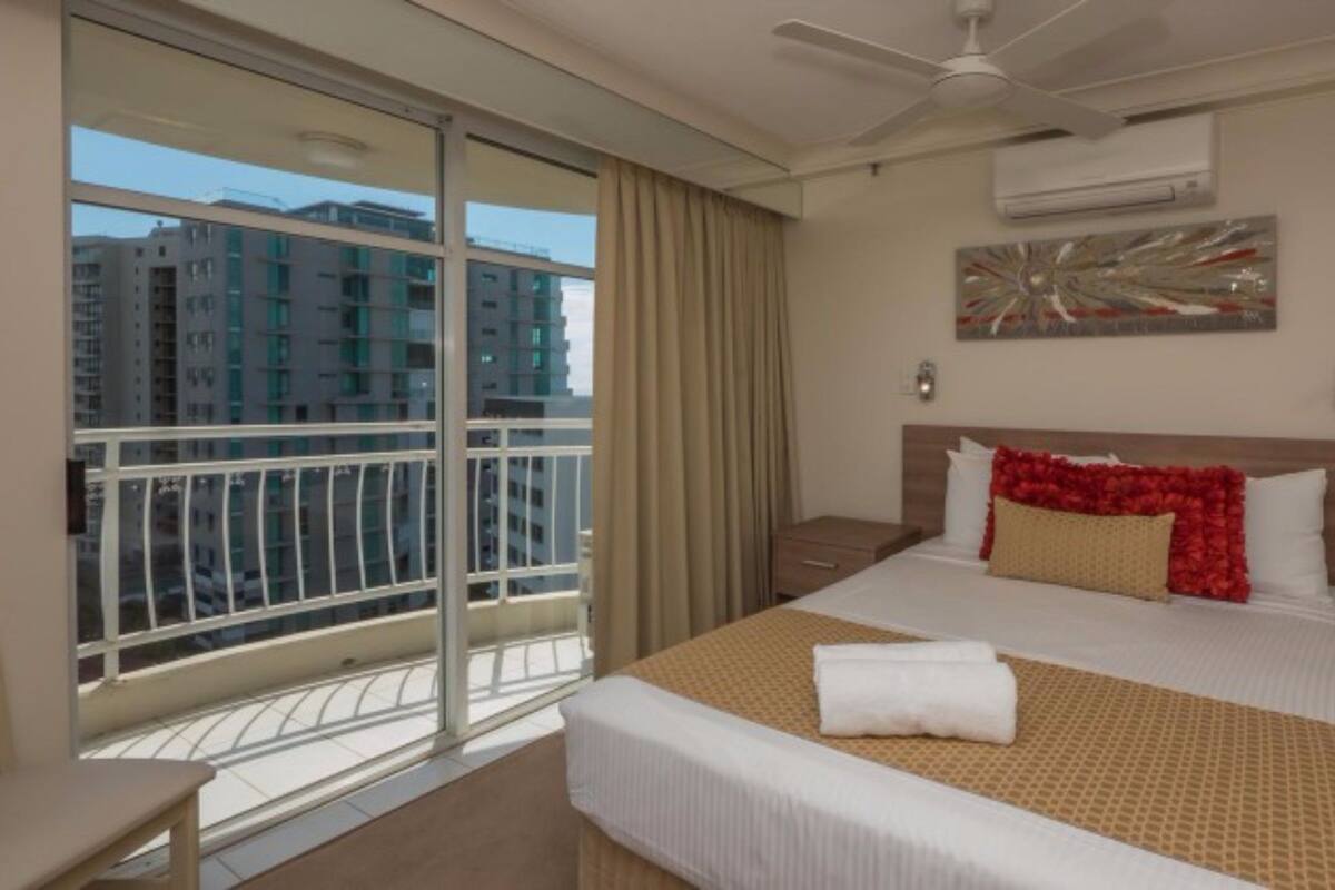 Une chambre d’hôtel avec un grand lit, un balcon donnant sur les bâtiments, un ventilateur de plafond et un appareil de climatisation.