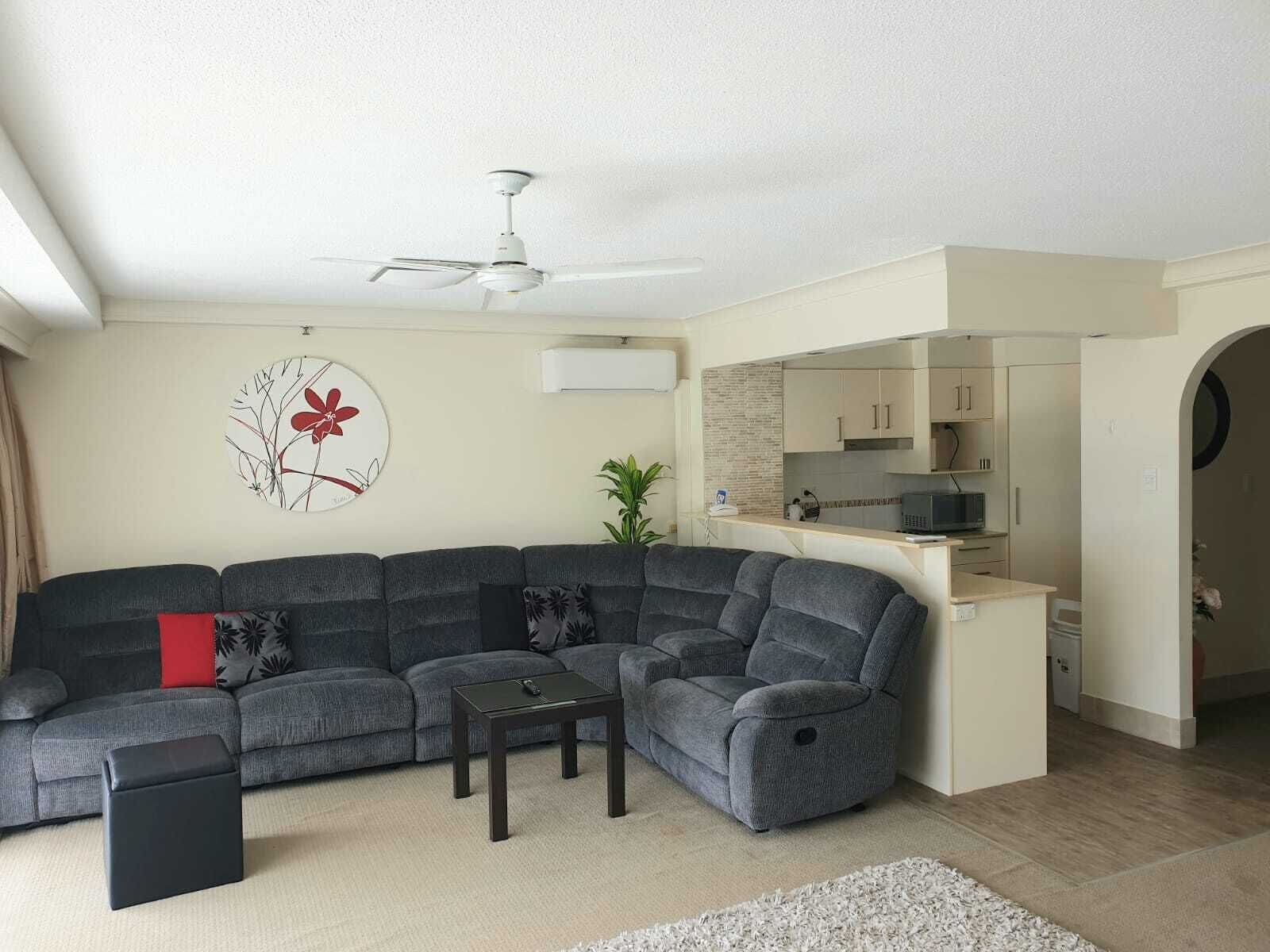 Una sala de estar moderna con un sofá seccional gris, un pequeño banquillo negro, una mesa de centro y un ventilador de techo. Se ve un área de cocina con armarios y un microondas a través de un umbral abierto.