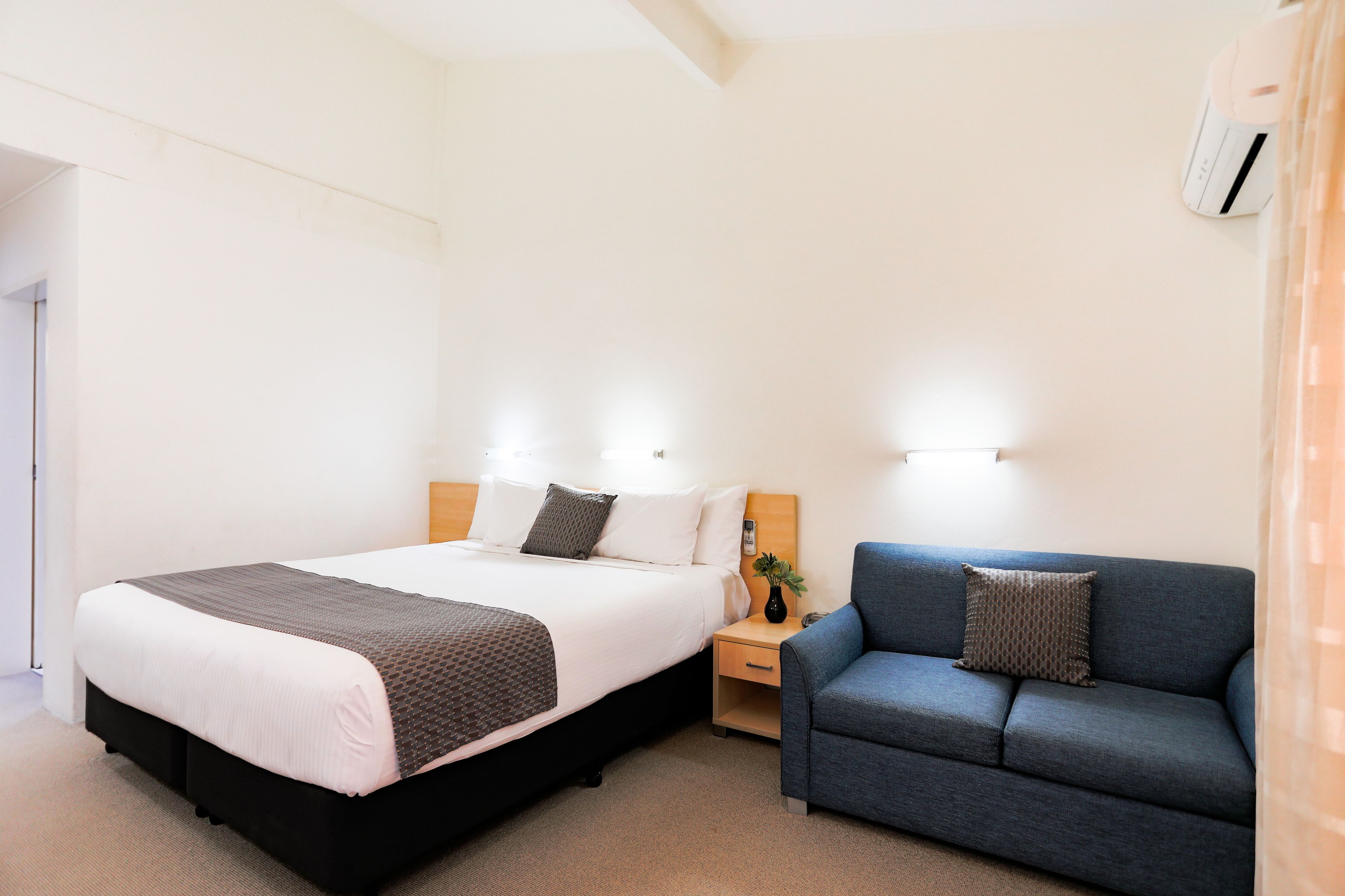 A hotel room with a bed, a nightstand, a blue sofa, and a wall-mounted air conditioner.