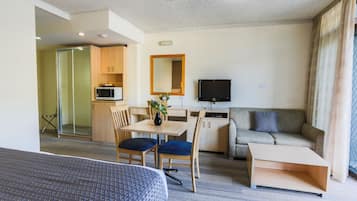 A modern hotel room with a bed, a dining table with chairs, a sofa, a TV, and a kitchenette.