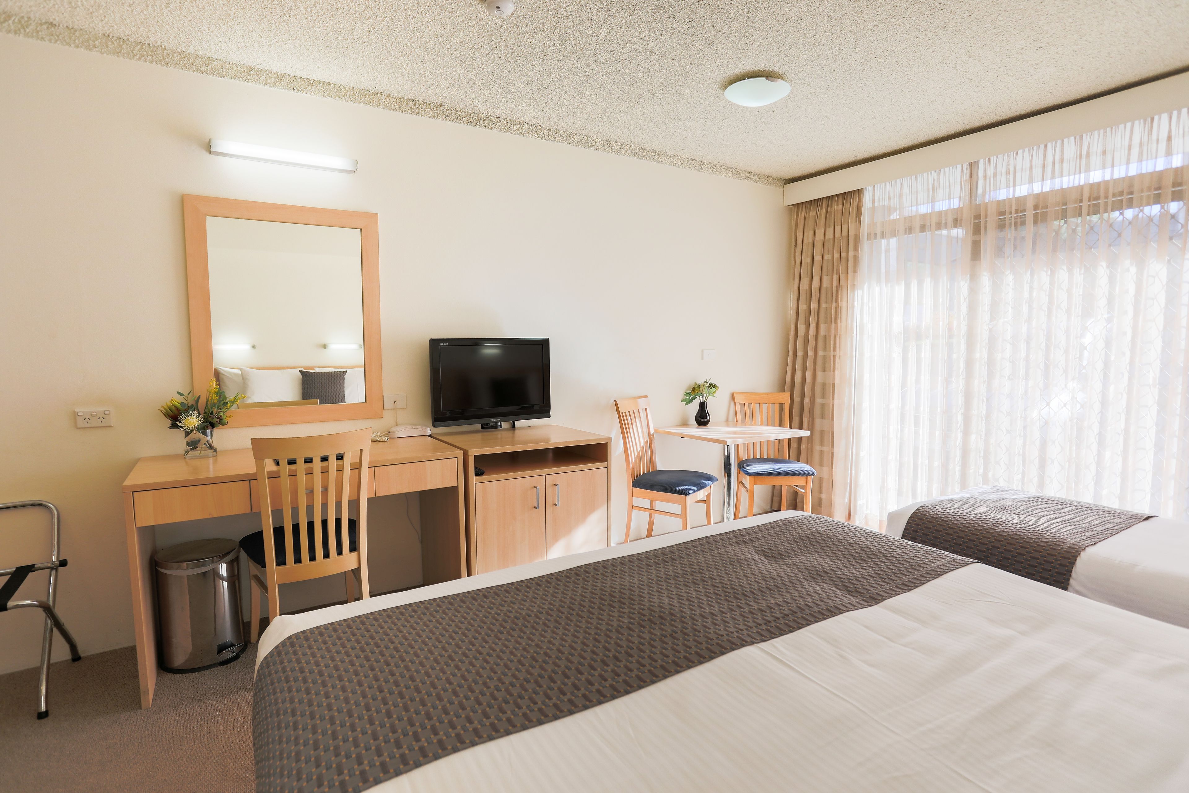 Une chambre d’hôtel avec deux lits, un bureau avec une télévision, une chaise, une petite table et une fenêtre avec des rideaux.