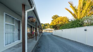 A building with a covered outdoor seating area, a paved pathway, and a white wall with a palm tree in the background. - Sierra Beachfront Motel (Kaikoura)