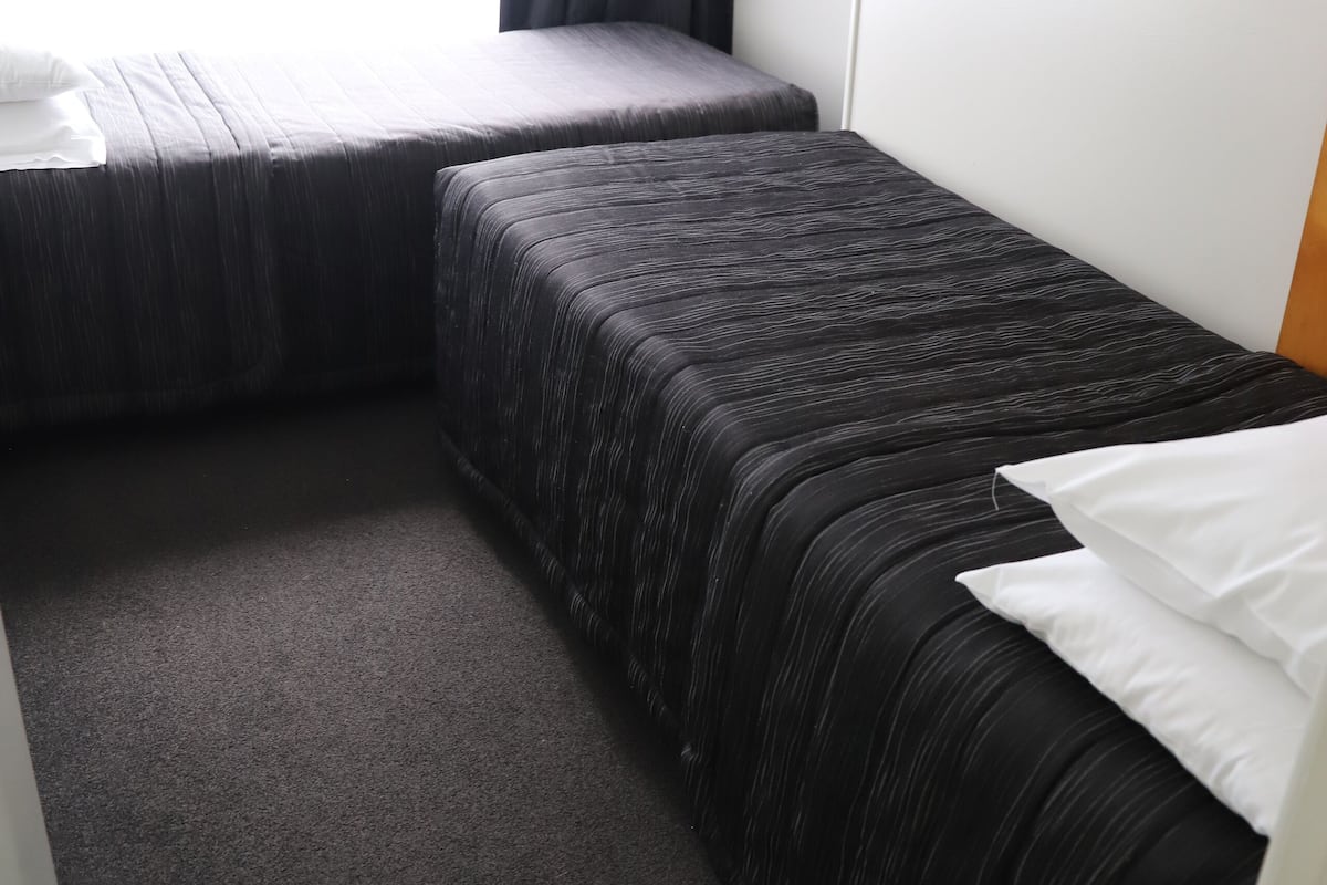 A small, compact room with two beds, a window with curtains, and a carpeted floor.