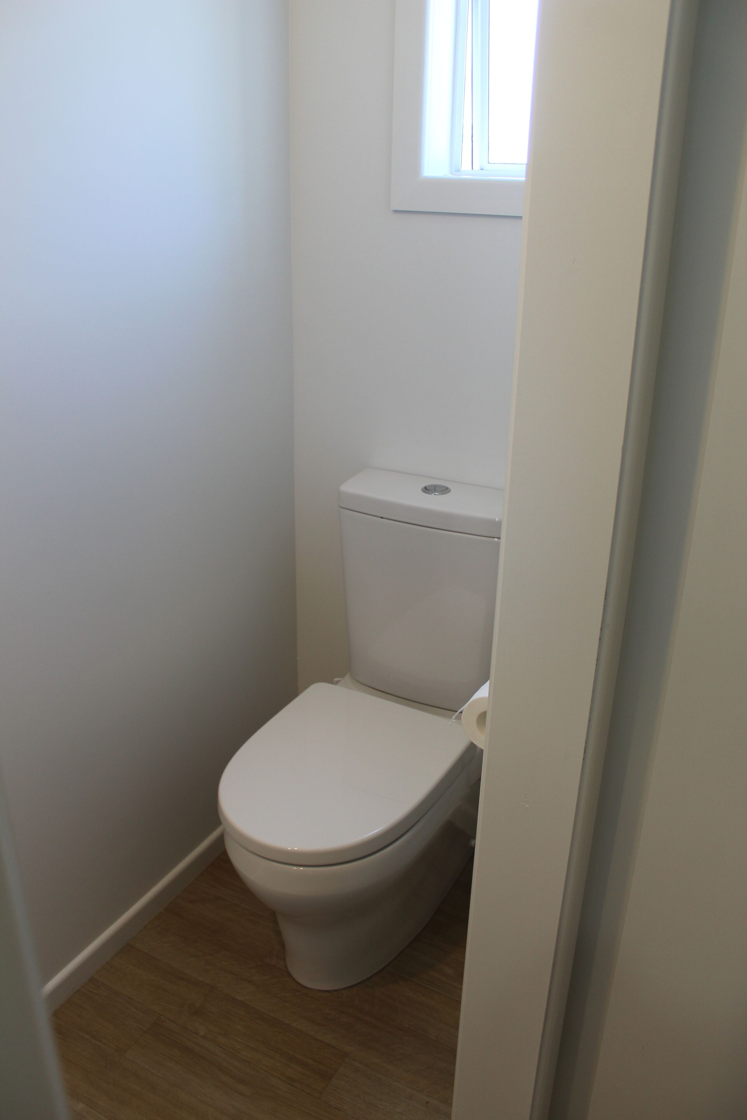 A small, white, single-occupancy bathroom with a toilet, a window above it, and wooden flooring.