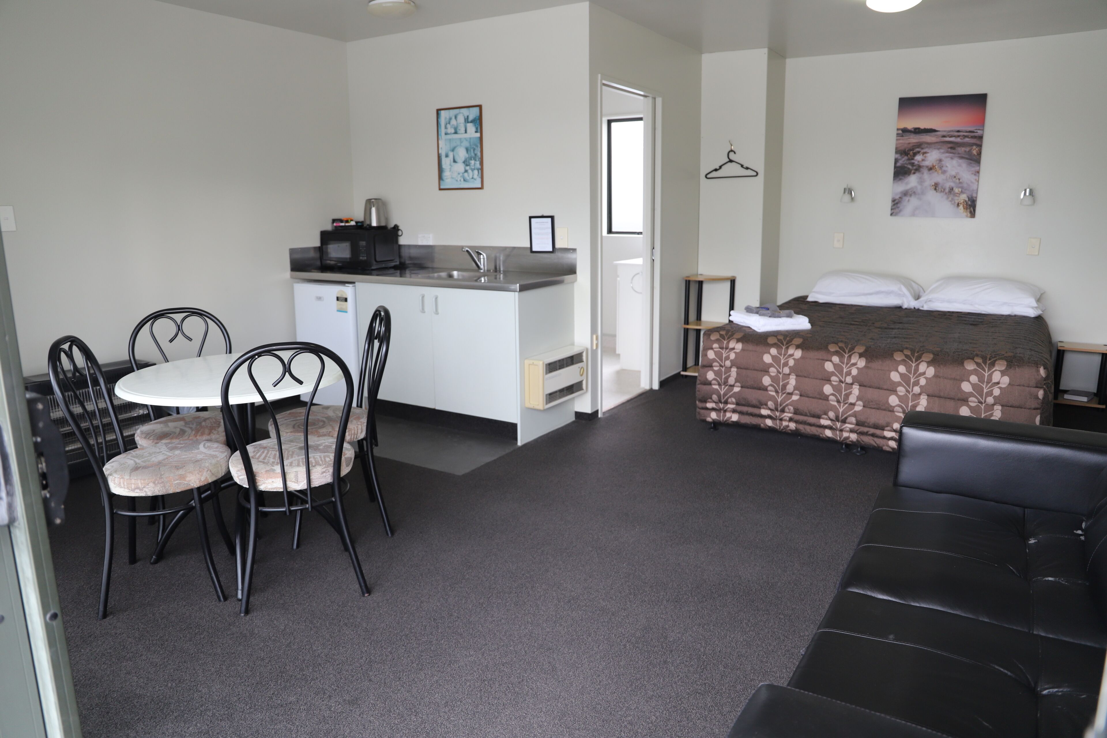 Ein kompaktes Zimmer mit einer Küchenzeile, einem Essbereich mit Stühlen, einem Bett mit gemustertem Überwurf und einem schwarzen Ledersofa.