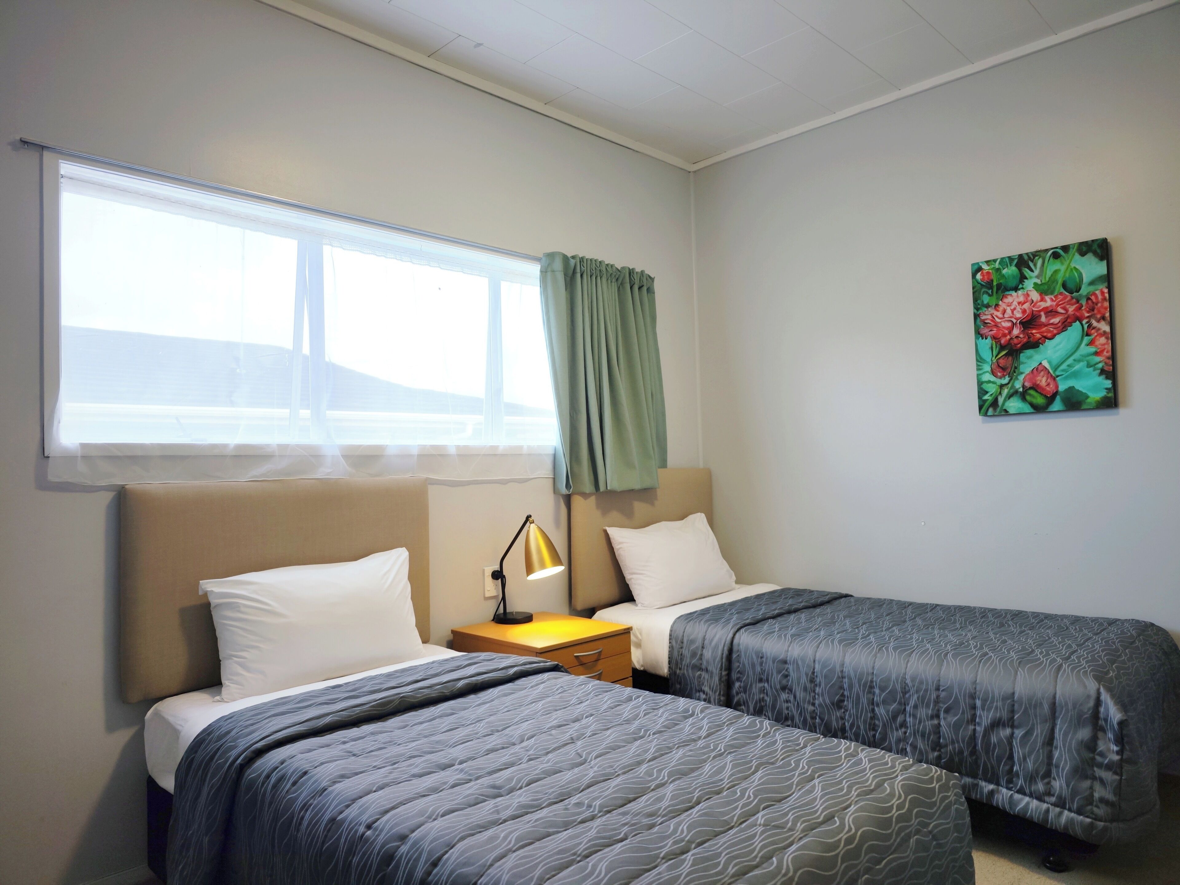 Hotellihuone, jossa on kaksi sänkyä, ikkuna, jossa on verhot, yöpöytä lampun kanssa ja taulu seinällä.
