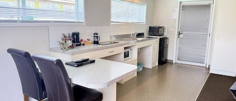 A modern kitchen with a central island, a microwave, a toaster, and a refrigerator. There is a dining area with two chairs and a table. An exit sign is visible on the wall.