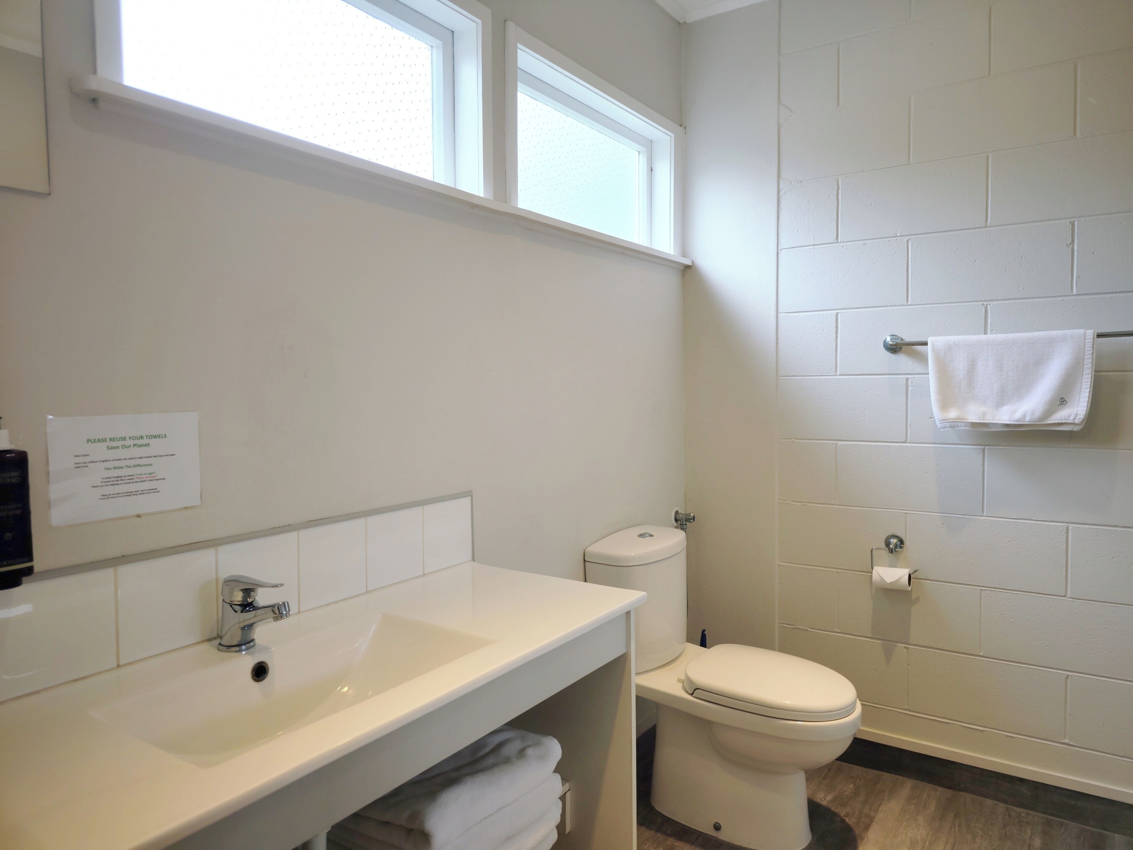 A bathroom with a white sink, toilet, and towel rack. There is a sign on the wall and a window above the sink.